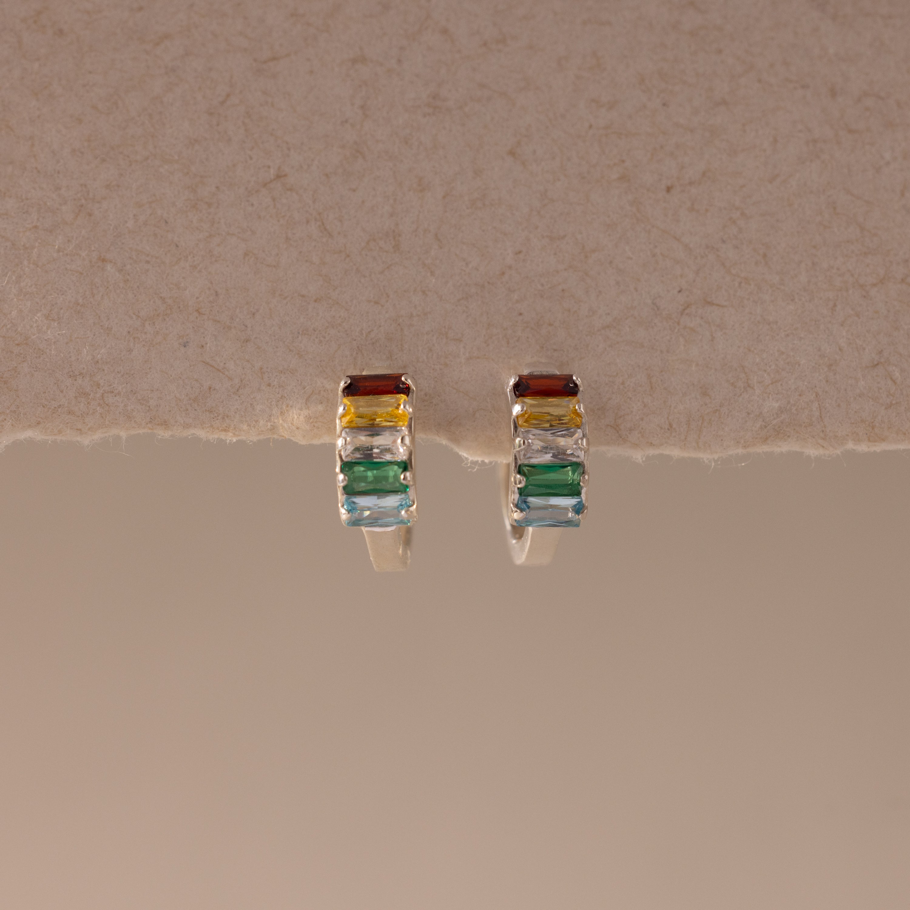 The Multiple Baguette Birthstone Huggies—silver petite hoops with rectangular red, yellow, blue, and green stones—rest on a textured beige surface, offering colorful jewelry for a playful accent to any look.
