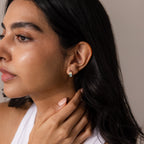 A woman with long dark hair, wearing a white top and petite Multiple Baguette Birthstone Huggies, gently touches her neck, adding a subtle pop of color to her look.