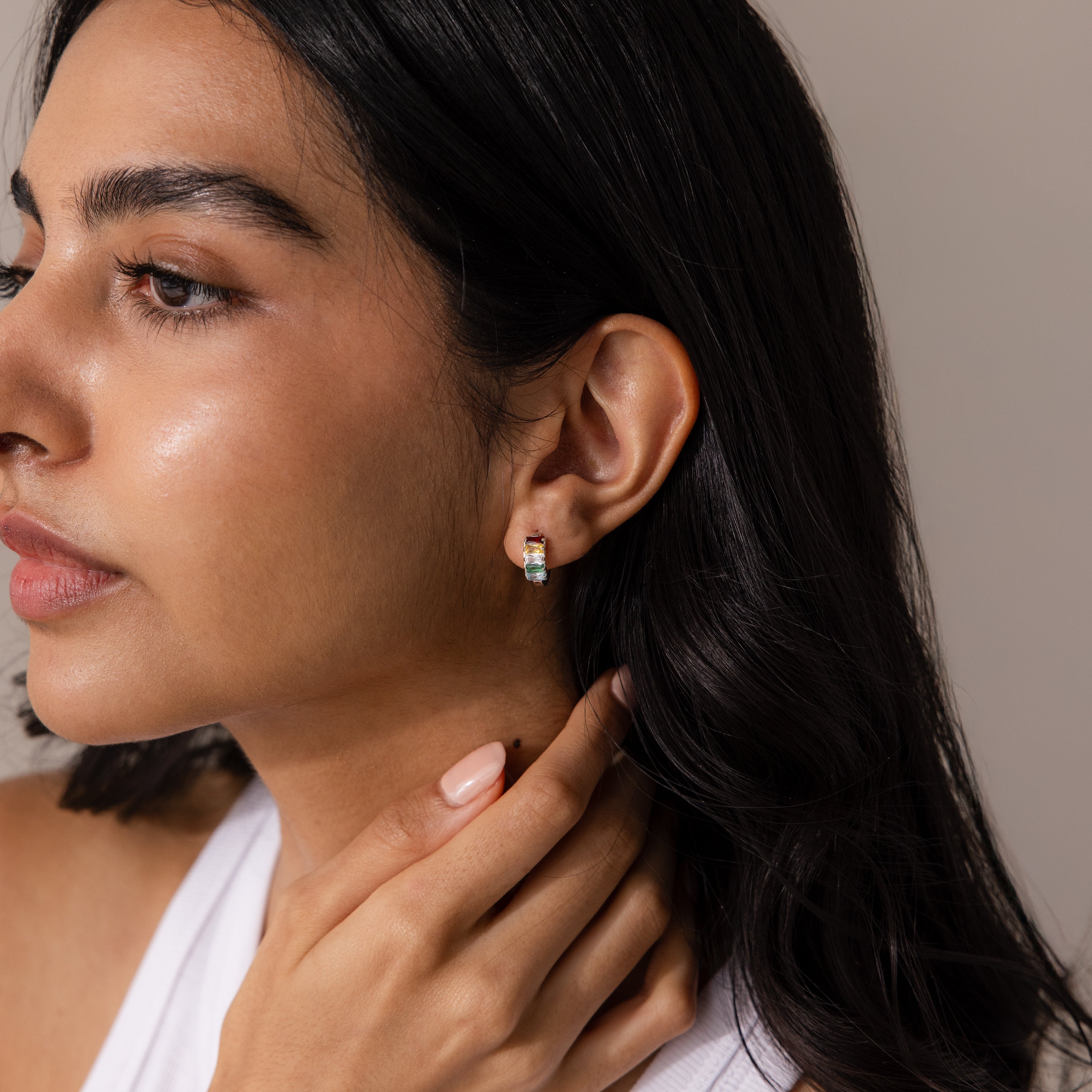 A woman with long dark hair, wearing a white top and petite Multiple Baguette Birthstone Huggies, gently touches her neck, adding a subtle pop of color to her look.