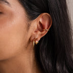 Close-up of a woman's ear wearing three gold hoop earrings, including petite Multiple Baguette Birthstone Huggies, with straight dark hair.
