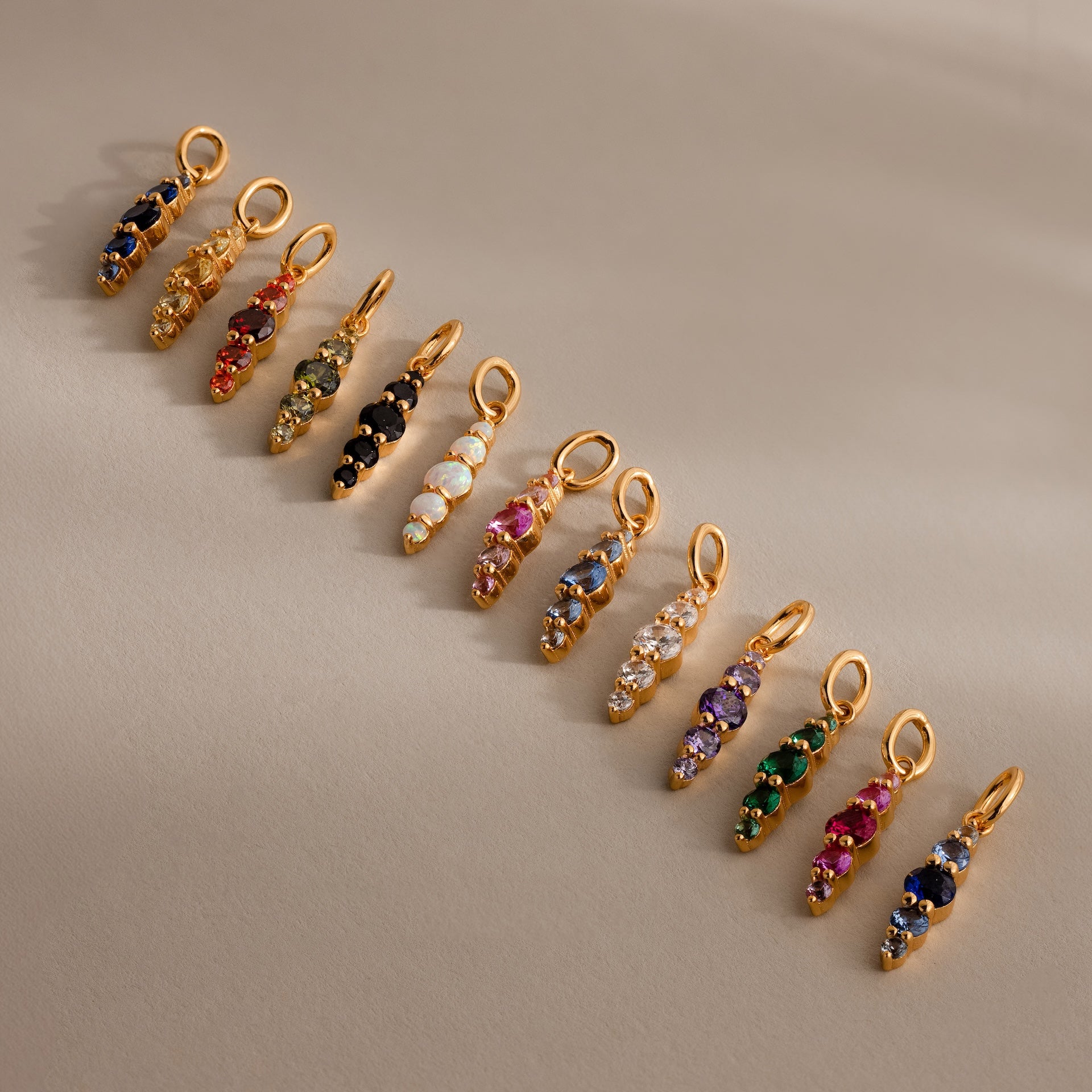 Twelve Ombre Lace Ruby Charm pendants, each with vibrant gemstone colors like birthstones, are neatly arranged on a beige surface.