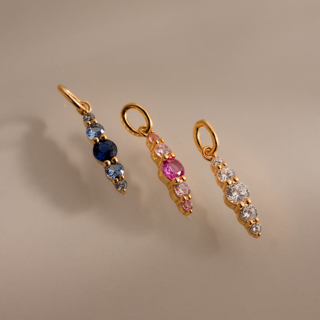 Three Ombre Lace Birthstone Charms—gold pendants with blue, pink, and clear gemstones—arranged vertically on a neutral background.