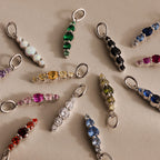 The Ombre Lace Birthstone Charm and other colorful gemstone charms are arranged in a circle on a light beige surface.