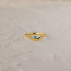 The Blue Zircon Drop Curve Ring, a gold stacking ring with a small blue gemstone, is displayed on a textured white surface.
