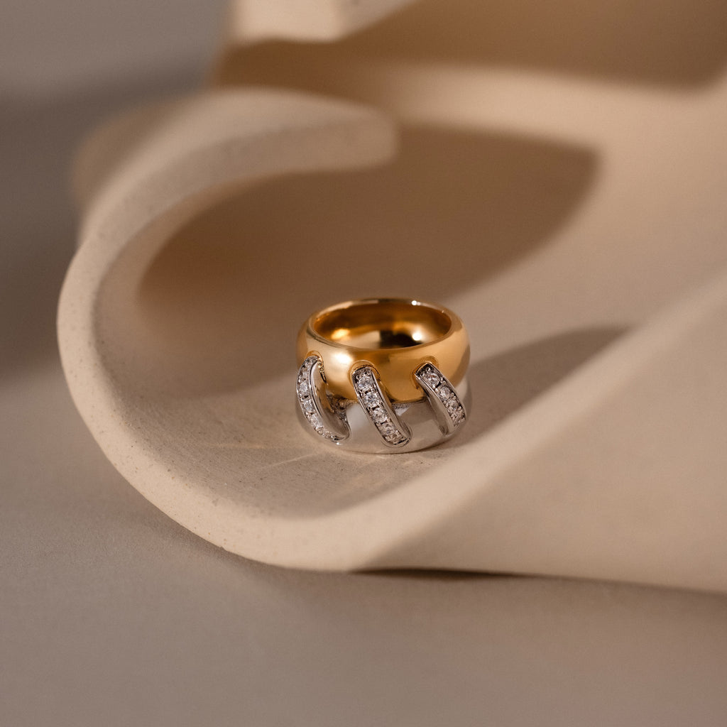 The Sculpted Mixed Metal Link Ring showcases gold and silver links accented with pavé diamonds, elegantly presented on a curved beige surface.