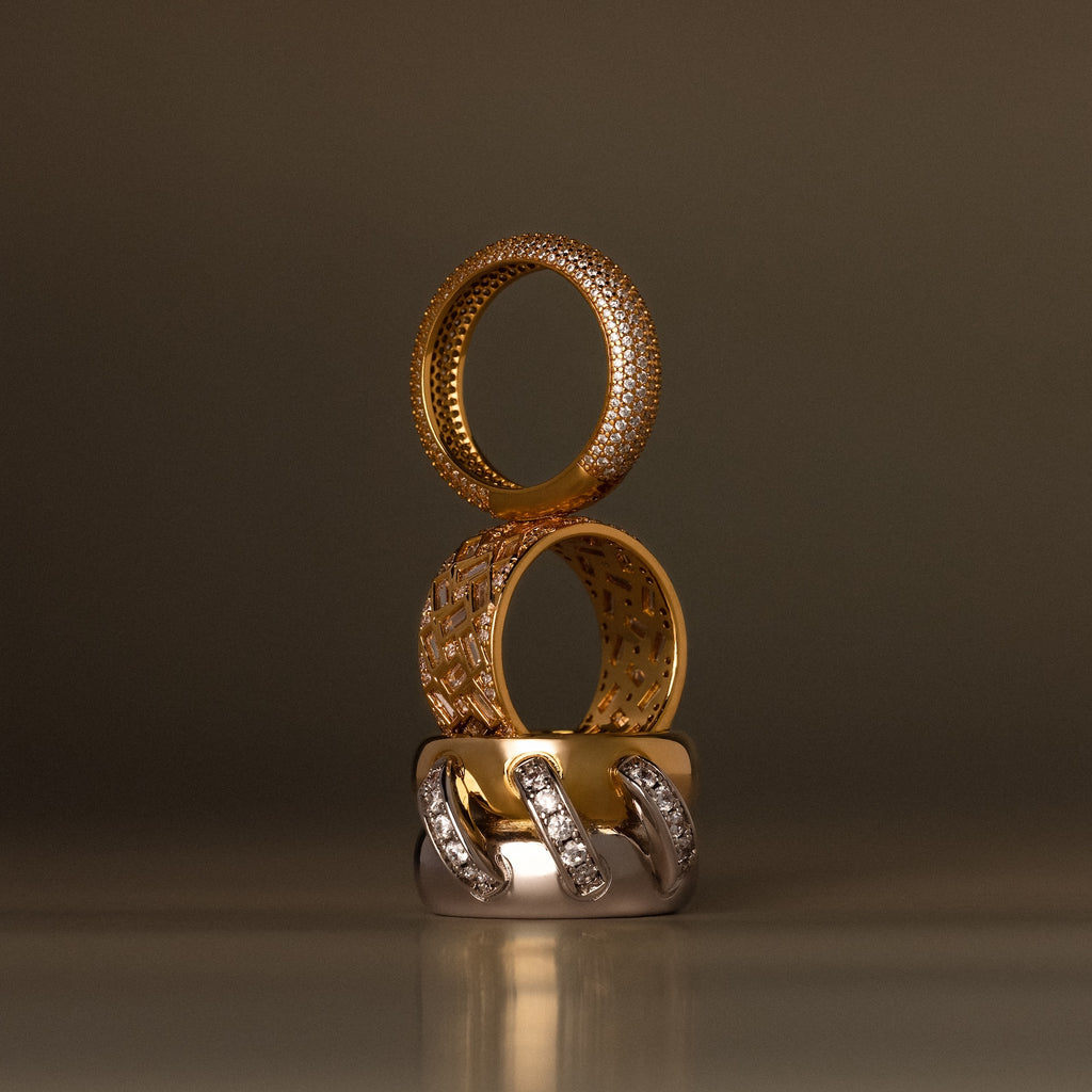 Three elegant rings, featuring the Mosaic Diamond Baguette Ring with baguette-cut stones, are stacked vertically in gold and silver tones with diamond accents against a neutral backdrop.