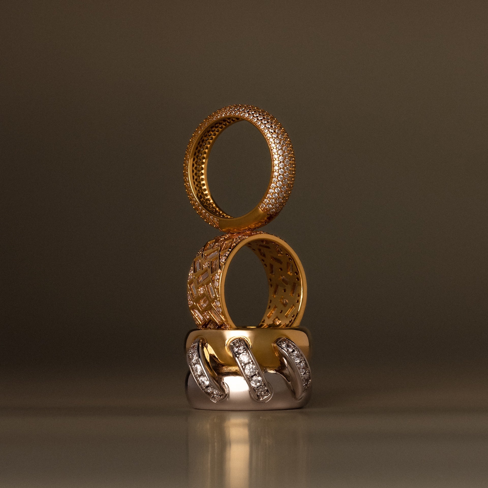 Three elegant rings, featuring the Mosaic Diamond Baguette Ring with baguette-cut stones, are stacked vertically in gold and silver tones with diamond accents against a neutral backdrop.