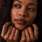 A woman with curly hair displays several gold stacking rings and the Dainty Ribbed Amethyst Ring on her fingers, gazing thoughtfully to the side.
