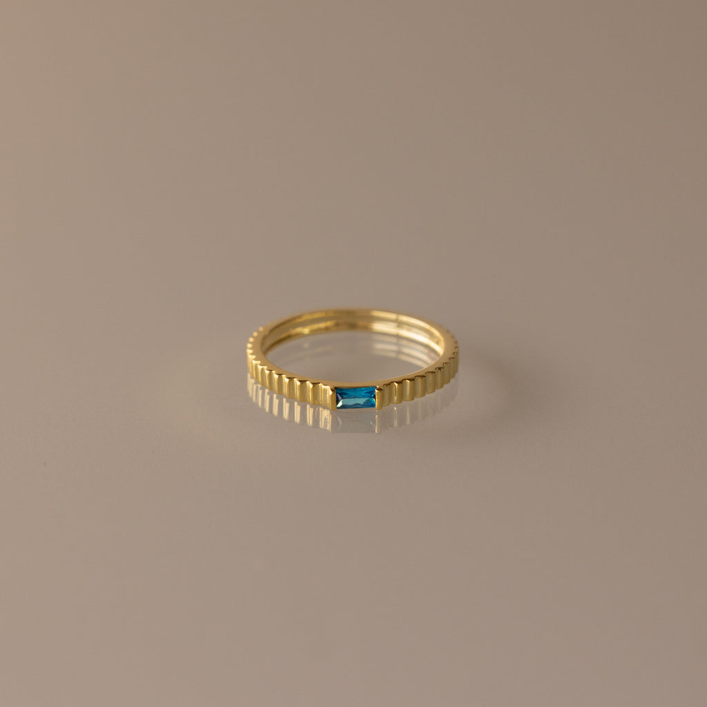 The Dainty Ribbed Blue Zircon Ring features a textured band and a small rectangular blue zircon gemstone, ideal as a birthstone piece or for stacking. Shown on a neutral background.