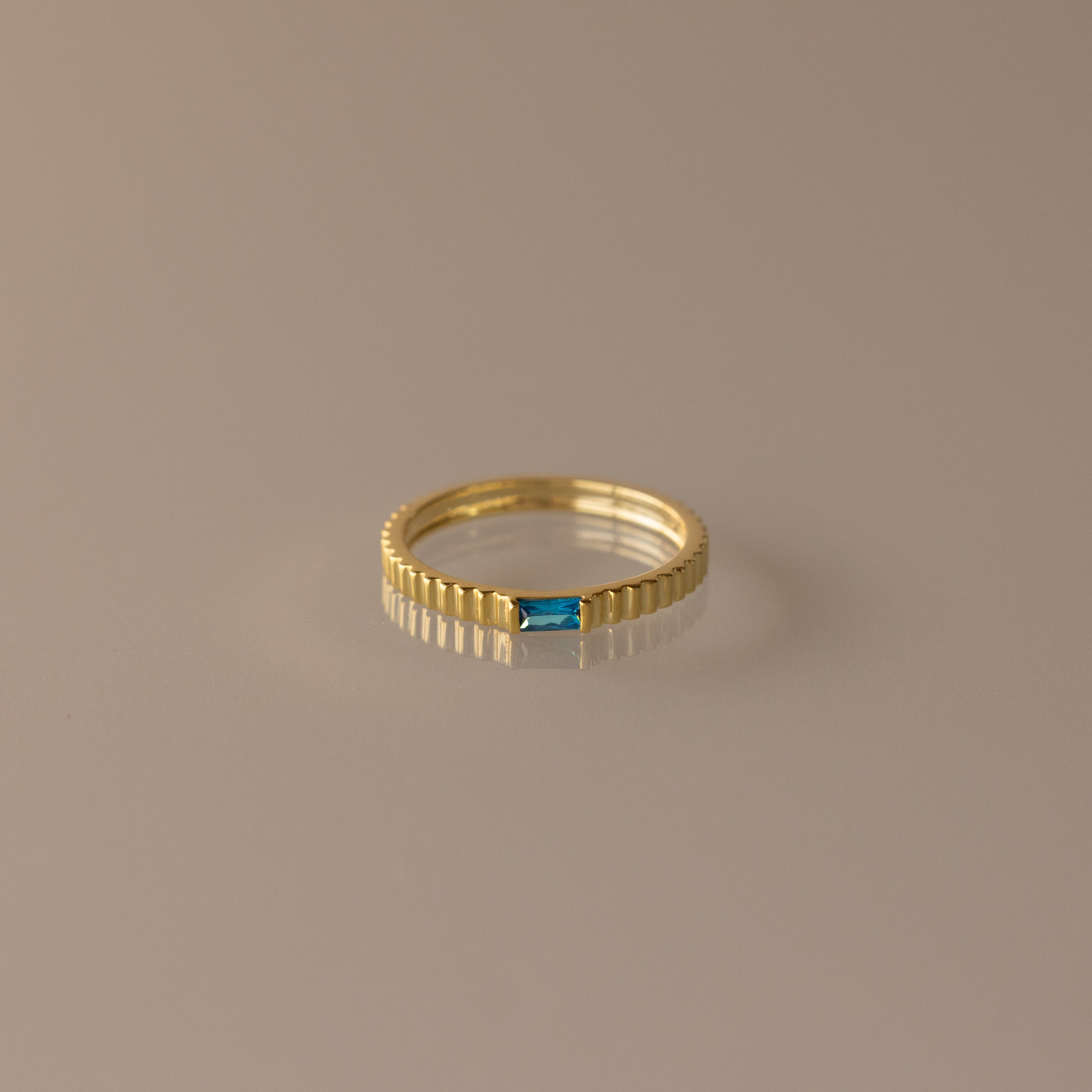 The Dainty Ribbed Blue Zircon Ring features a textured band and a small rectangular blue zircon gemstone, ideal as a birthstone piece or for stacking. Shown on a neutral background.