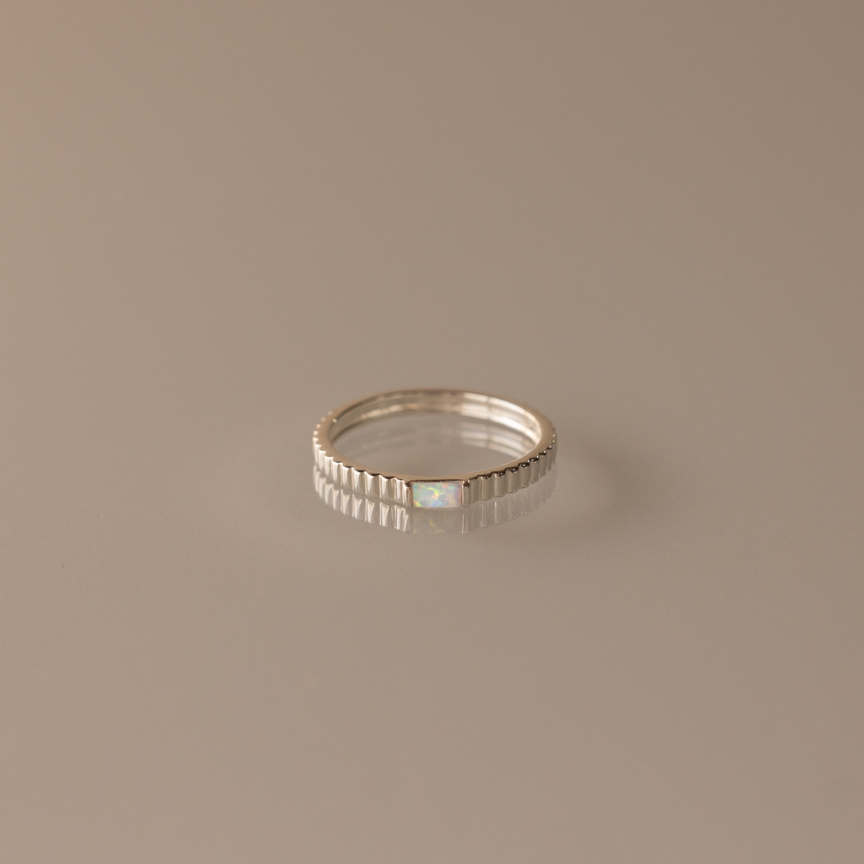 The Dainty Ribbed Opal Ring features a silver ribbed band with a small, subtle opal at the center, showcased on a beige background.
