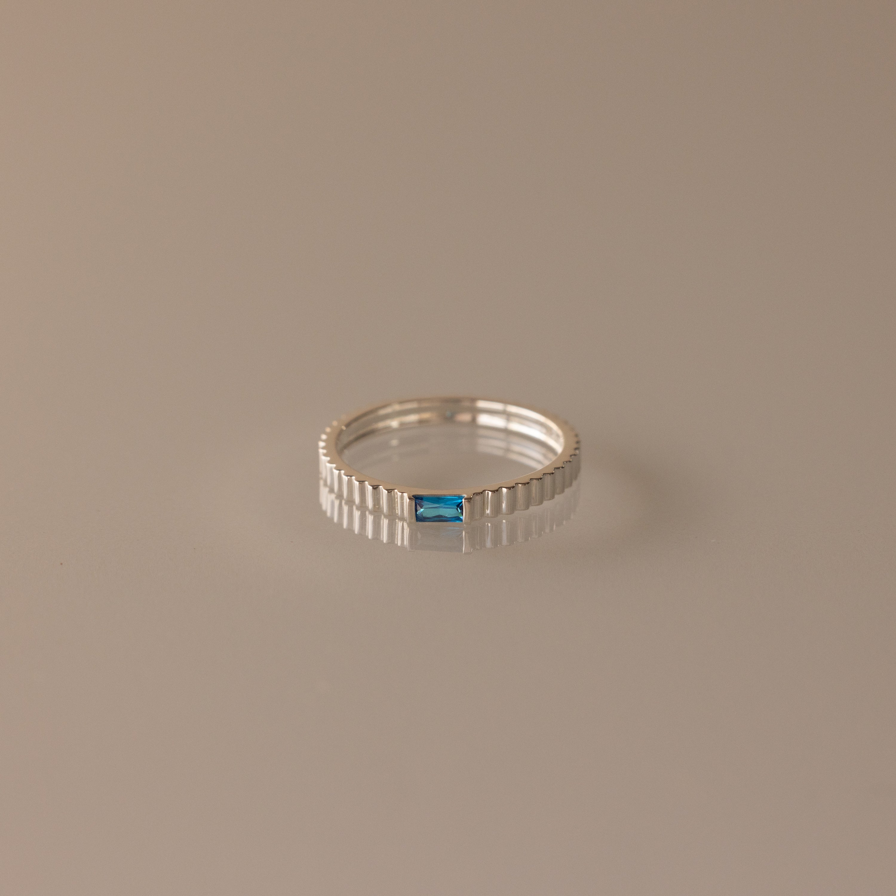 The Dainty Ribbed Blue Zircon Ring showcases a rectangular blue zircon gemstone at the center, set in a delicate silver band and elegantly displayed on a neutral background.