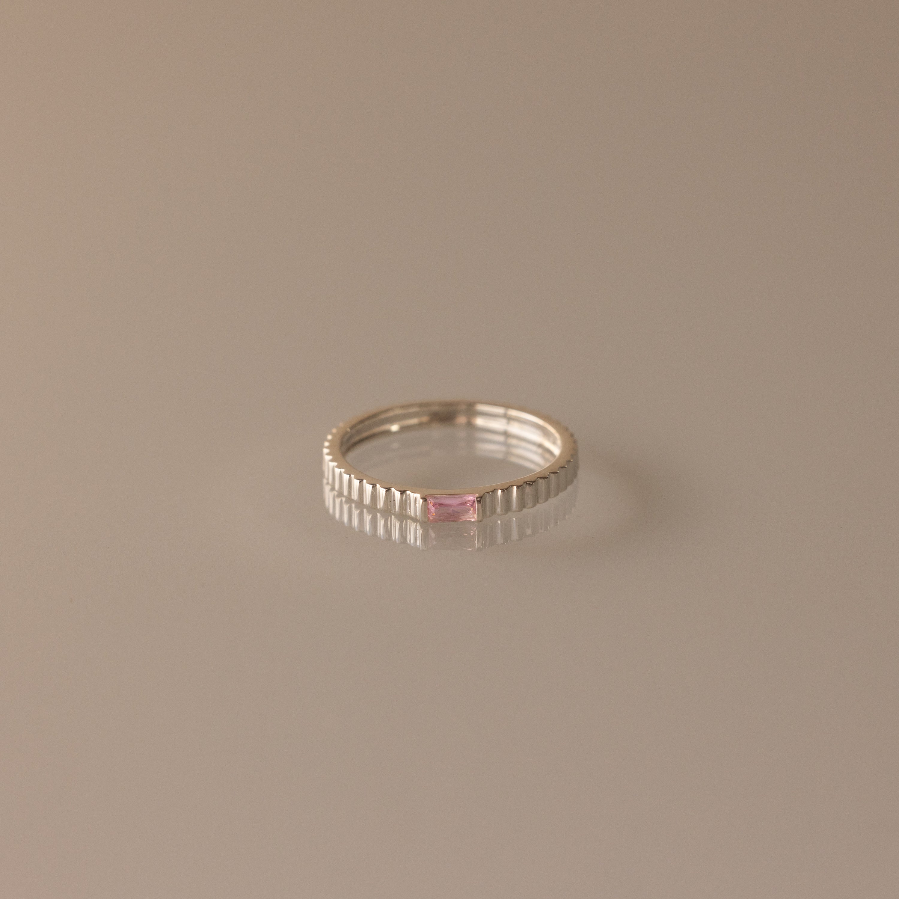 The Dainty Ribbed Pink Tourmaline Ring showcases a pink tourmaline centerpiece set on a sleek silver ribbed band, displayed on a smooth beige surface.
