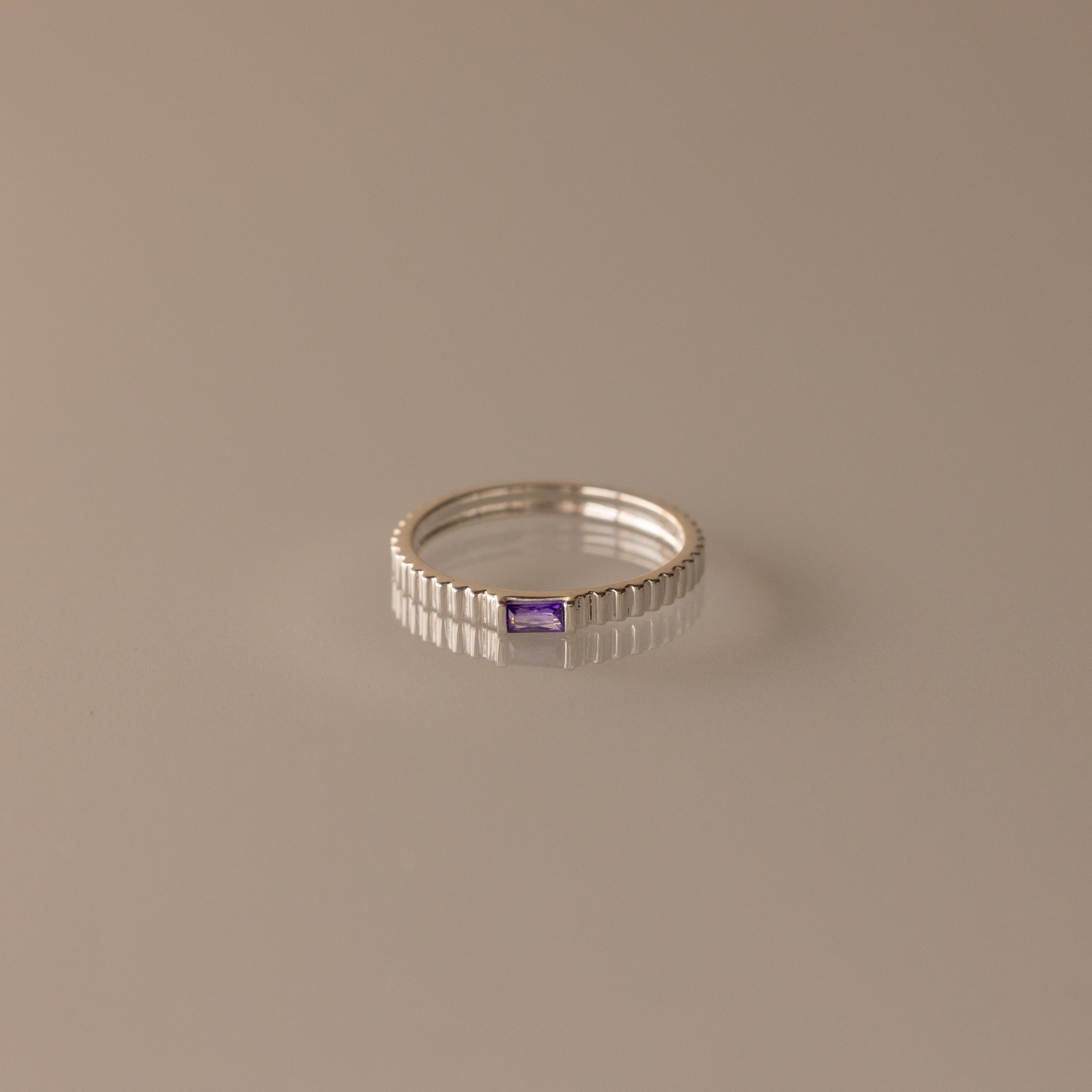 The Dainty Ribbed Amethyst Ring features a rectangular, ribbed band and a small center amethyst gemstone, shown on a beige background.