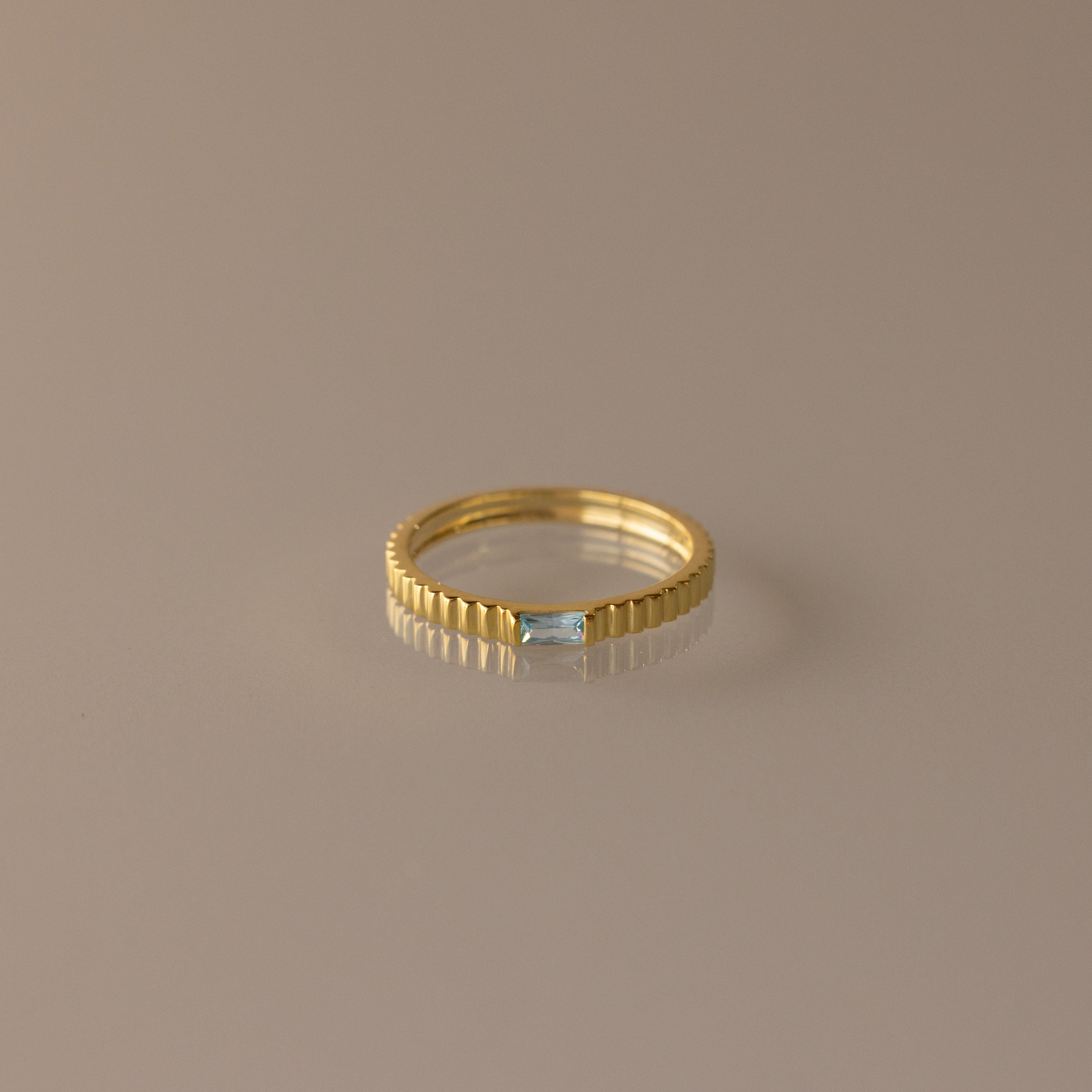 The Dainty Ribbed Aquamarine Ring features a gold band with a rectangular aquamarine gemstone at its center, shown against a neutral background.
