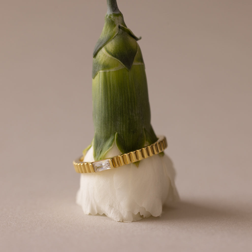 The Dainty Ribbed Topaz Ring, featuring a rectangular gemstone, is elegantly stacked around the stem of a white flower bud.