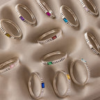 The Dainty Ribbed Peridot Ring is displayed neatly on a beige, textured surface alongside other silver gemstone rings.