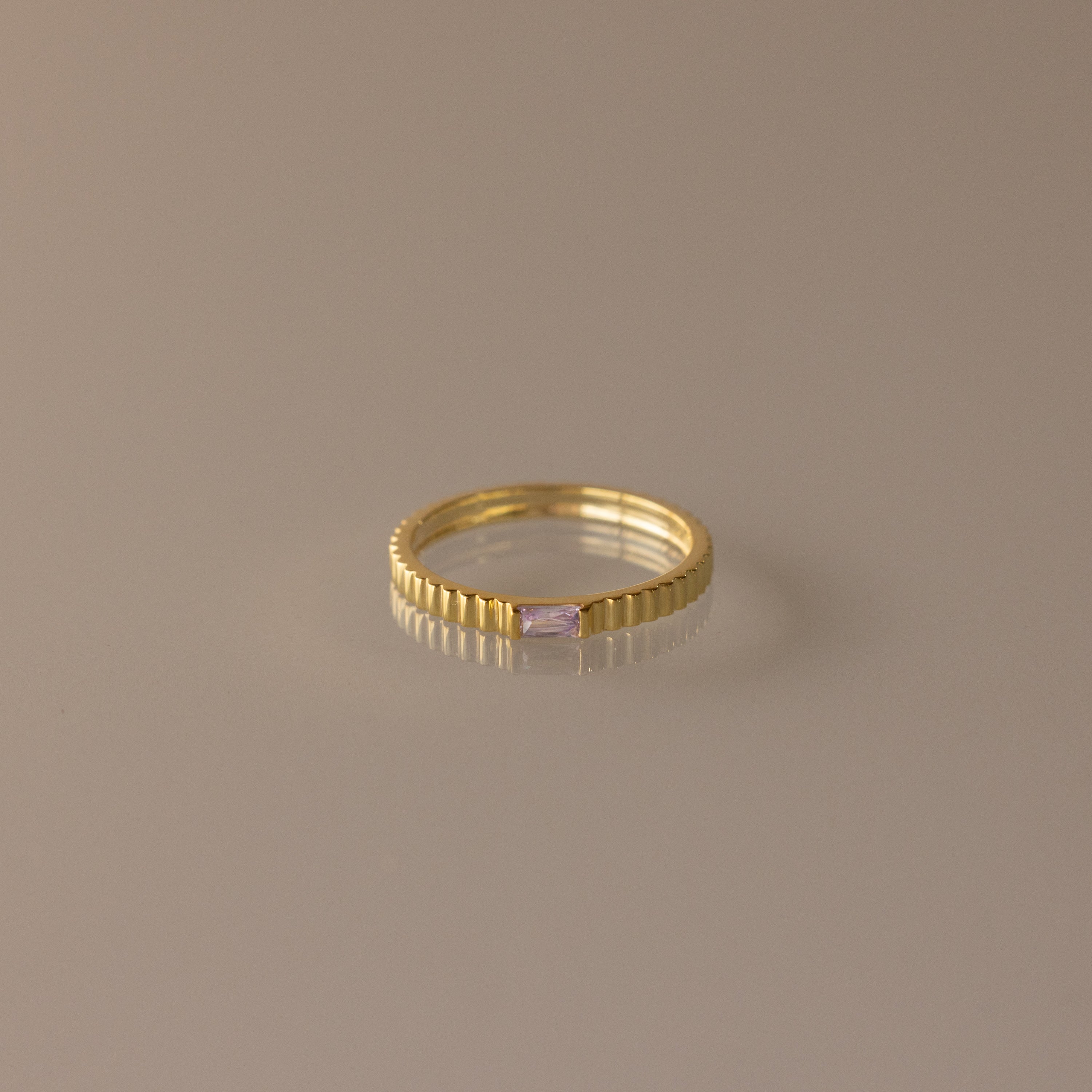 The Dainty Ribbed Alexandrite Ring features a gold ridged band and a small rectangular pale pink gemstone, ideal as a delicate ring or unique birthstone piece. Displayed on a plain surface.
