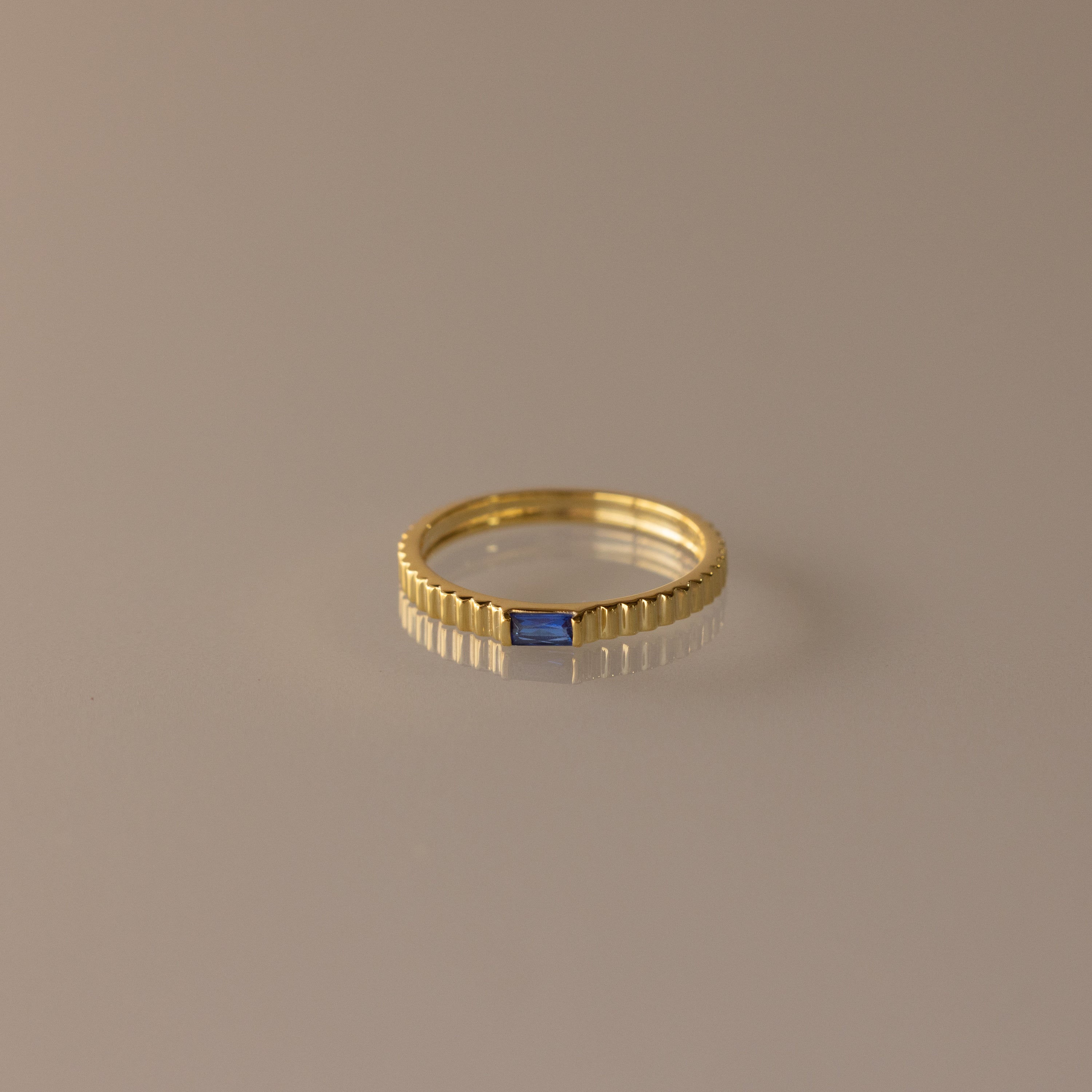 The Dainty Ribbed Sapphire Ring features a textured band and a petite rectangular blue sapphire, ideal for stacking and shown on a neutral background.