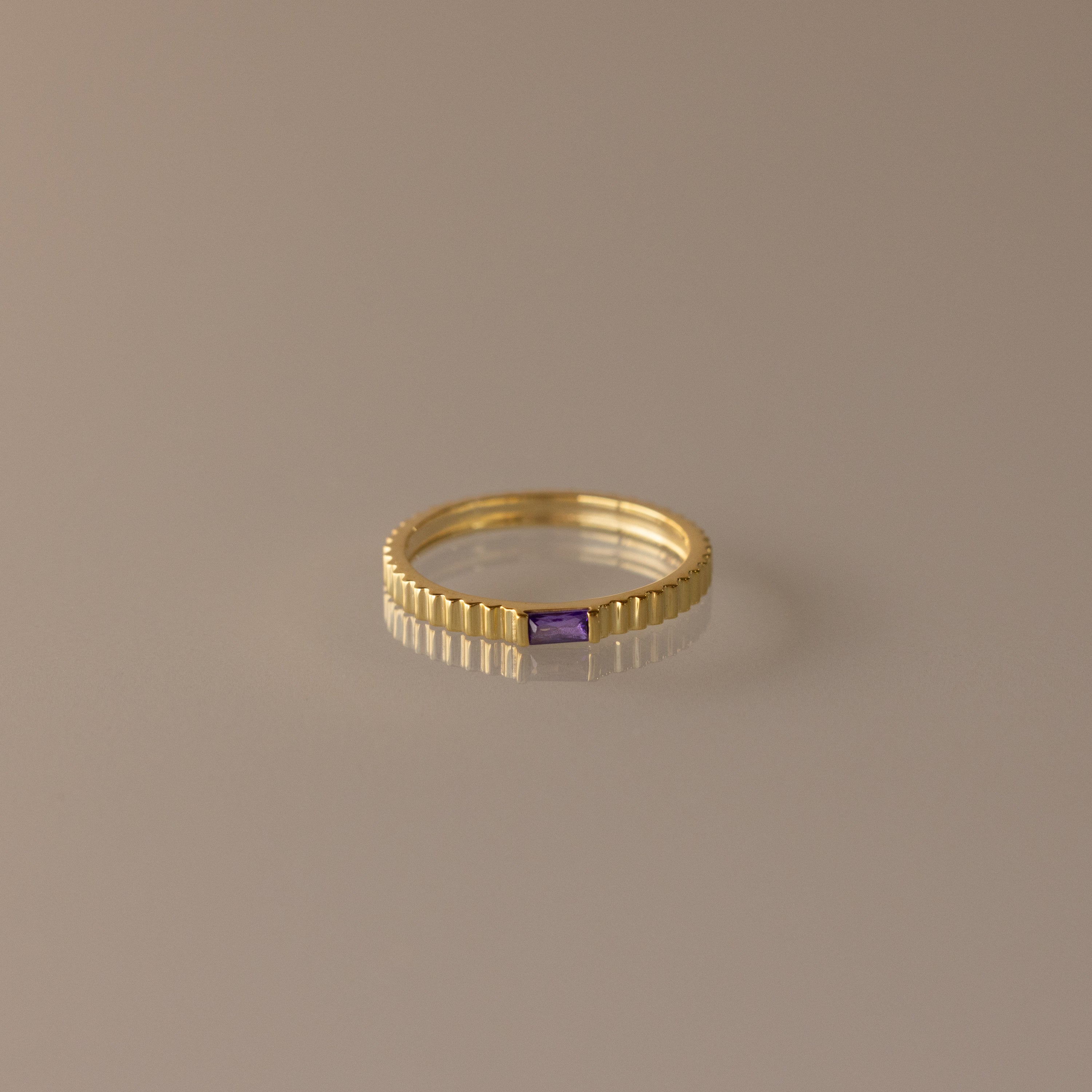 The Dainty Ribbed Amethyst Ring features a gold ribbed band and a small rectangular purple amethyst, perfect for stacking or as a delicate birthstone ring. Shown on a neutral background.