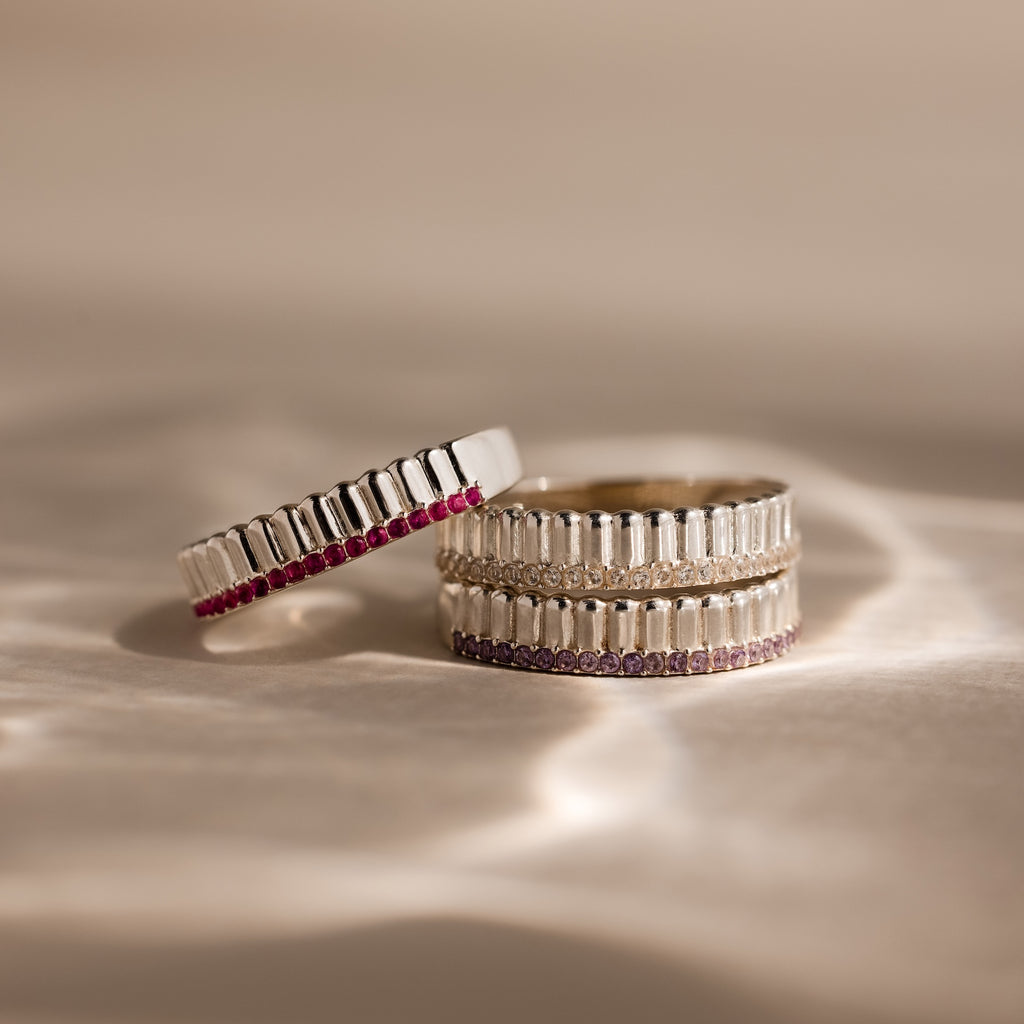 Three Ribbed Ruby Stack Rings, crafted in silver with vertical grooves and accented by small ruby stones, are displayed on a soft beige background.