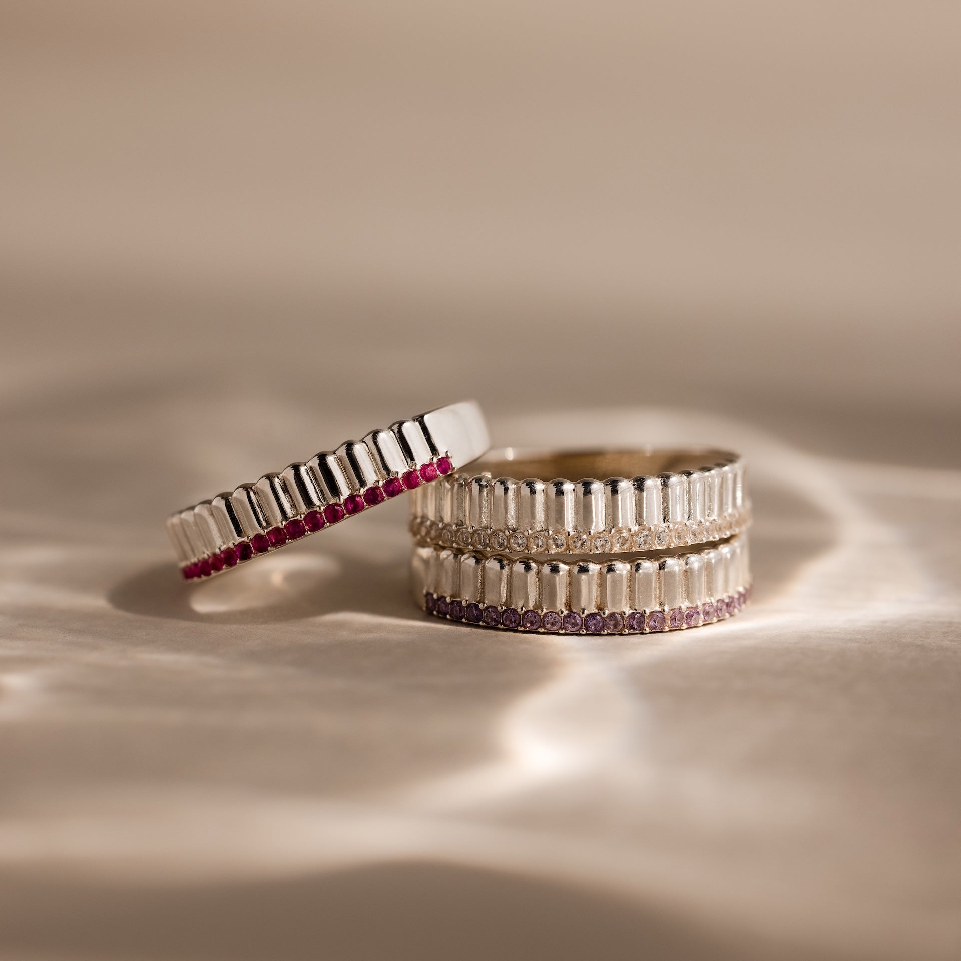 The Ribbed Pink Tourmaline Stack Ring features three silver bands with a sculpted ribbed texture; two stacked stylishly and one leaning, adorned with small pink and clear stones.
