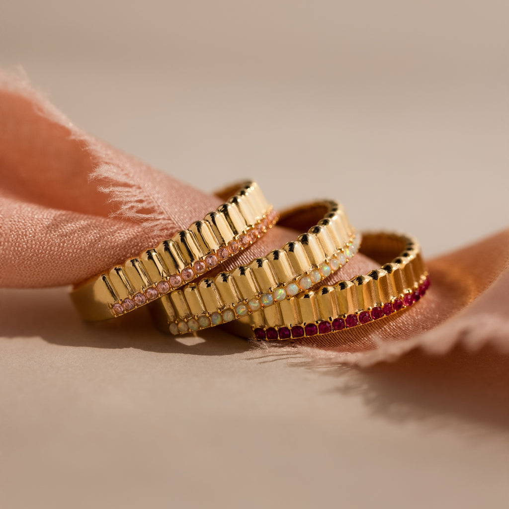Three gold rings with small colorful gemstones, including a Ribbed Ruby Stack Ring, are stacked on pink fabric with a soft beige background.