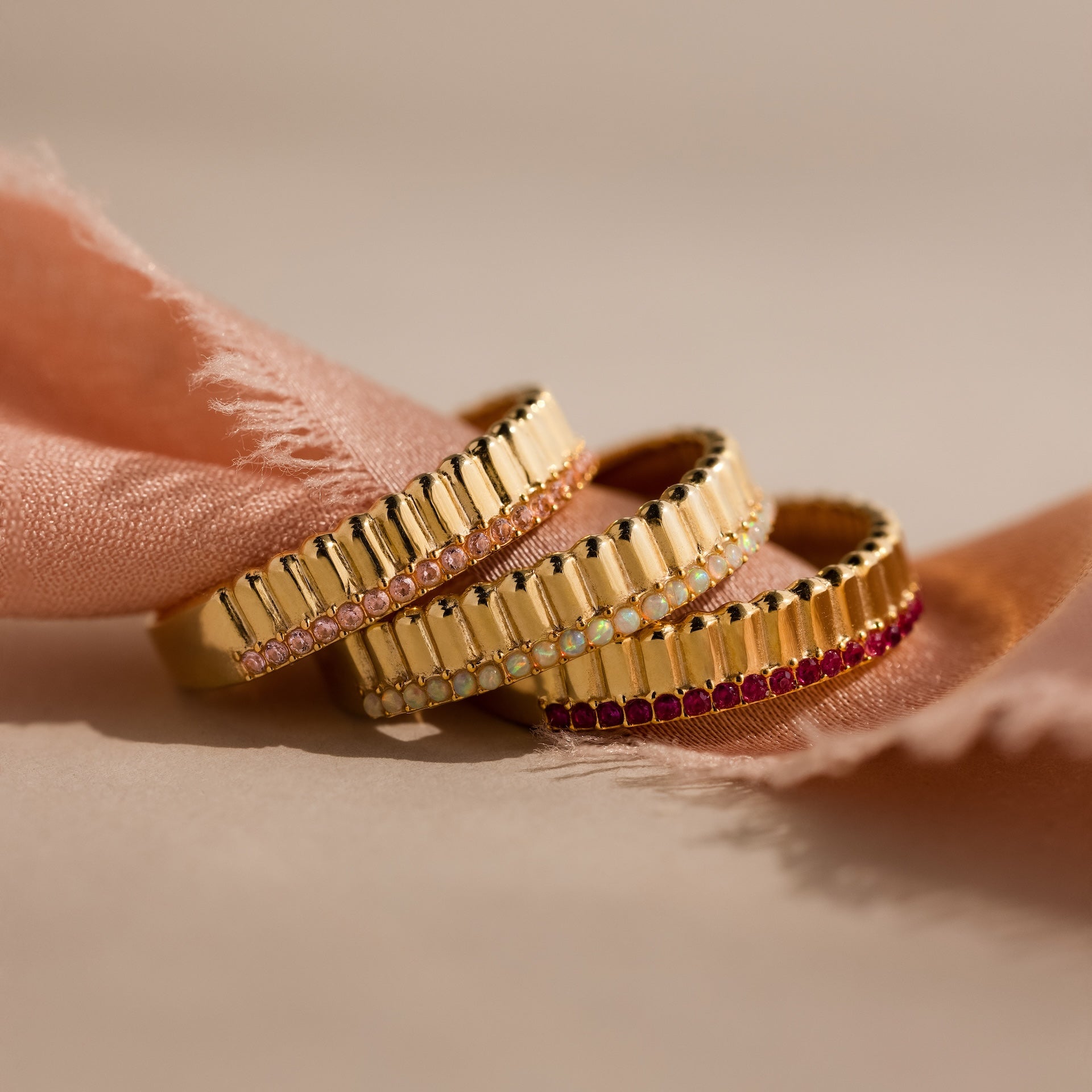 Three gold rings with small colorful gemstones, including a Ribbed Ruby Stack Ring, are stacked on pink fabric with a soft beige background.