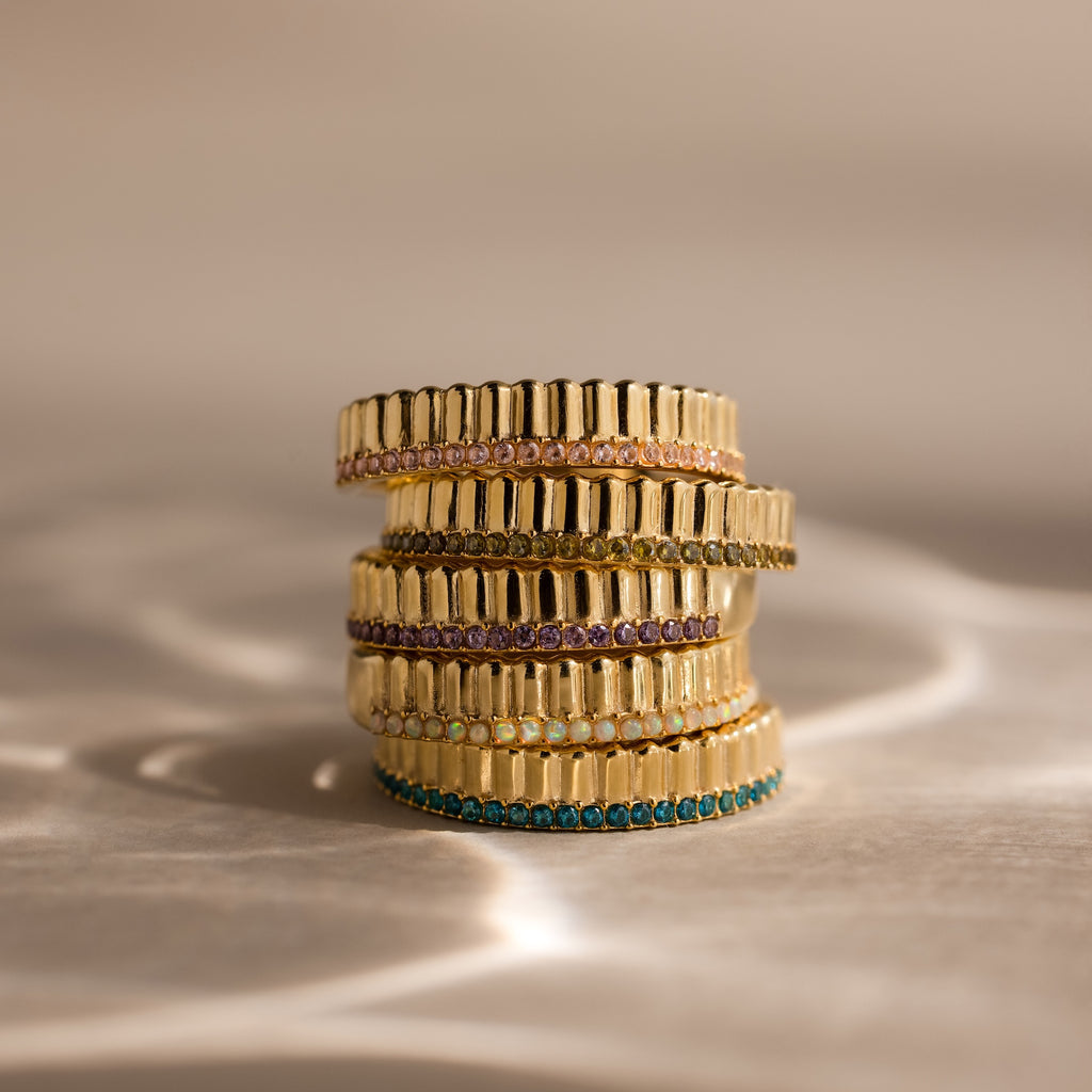 The Ribbed Sapphire Stack Ring features four stacked gold bands, each set with colorful sapphires and a sculpted ribbed texture, creating a modern yet timeless look against a reflective surface with a soft background.