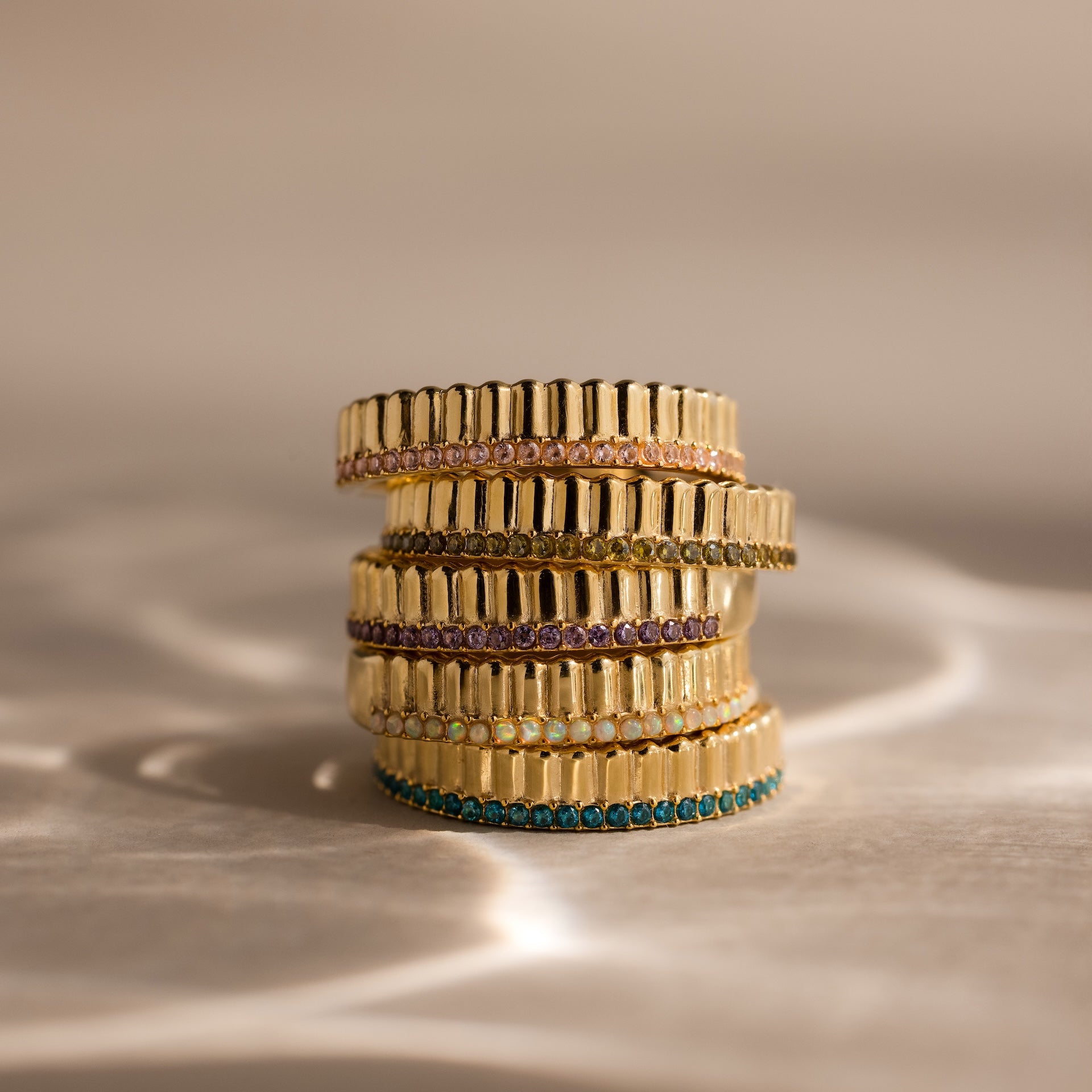 The Ribbed Emerald Stack Ring, featuring a sculpted ribbed texture and vibrant colored gemstone band, is showcased stacked on a reflective beige surface.