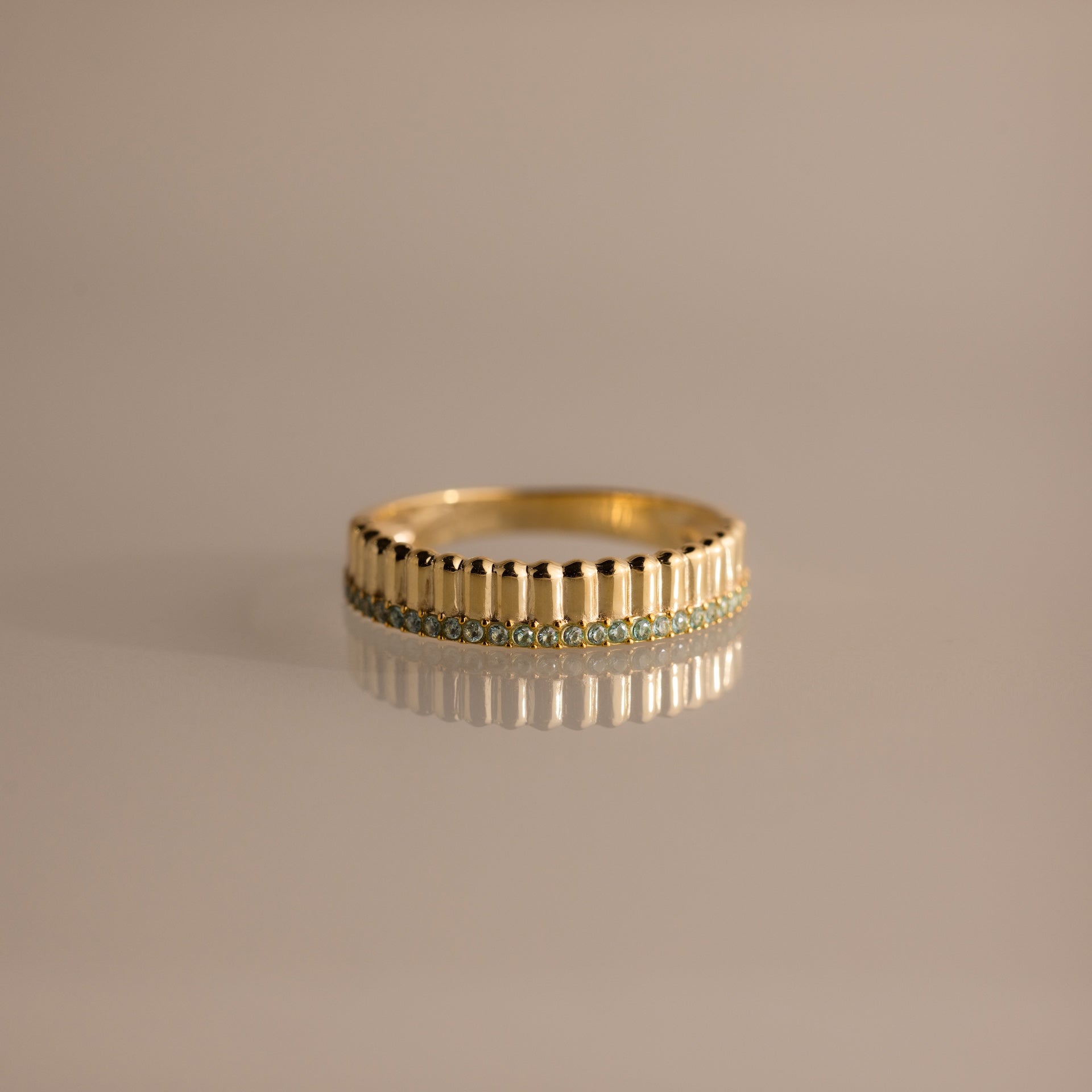 The Ribbed Aquamarine Stack Ring is a modern, stackable ring with vertical ribbed texture and a row of small aquamarine stones, shown on a reflective surface.
