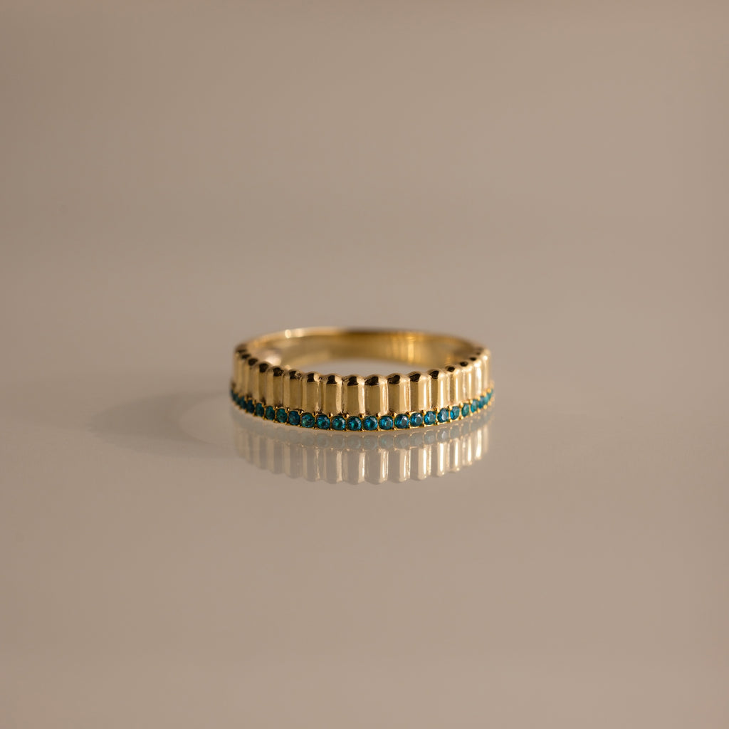 The Ribbed Blue Zircon Stack Ring features a gold band with small blue stones in a row, perfect for stacking and shown on a reflective surface.
