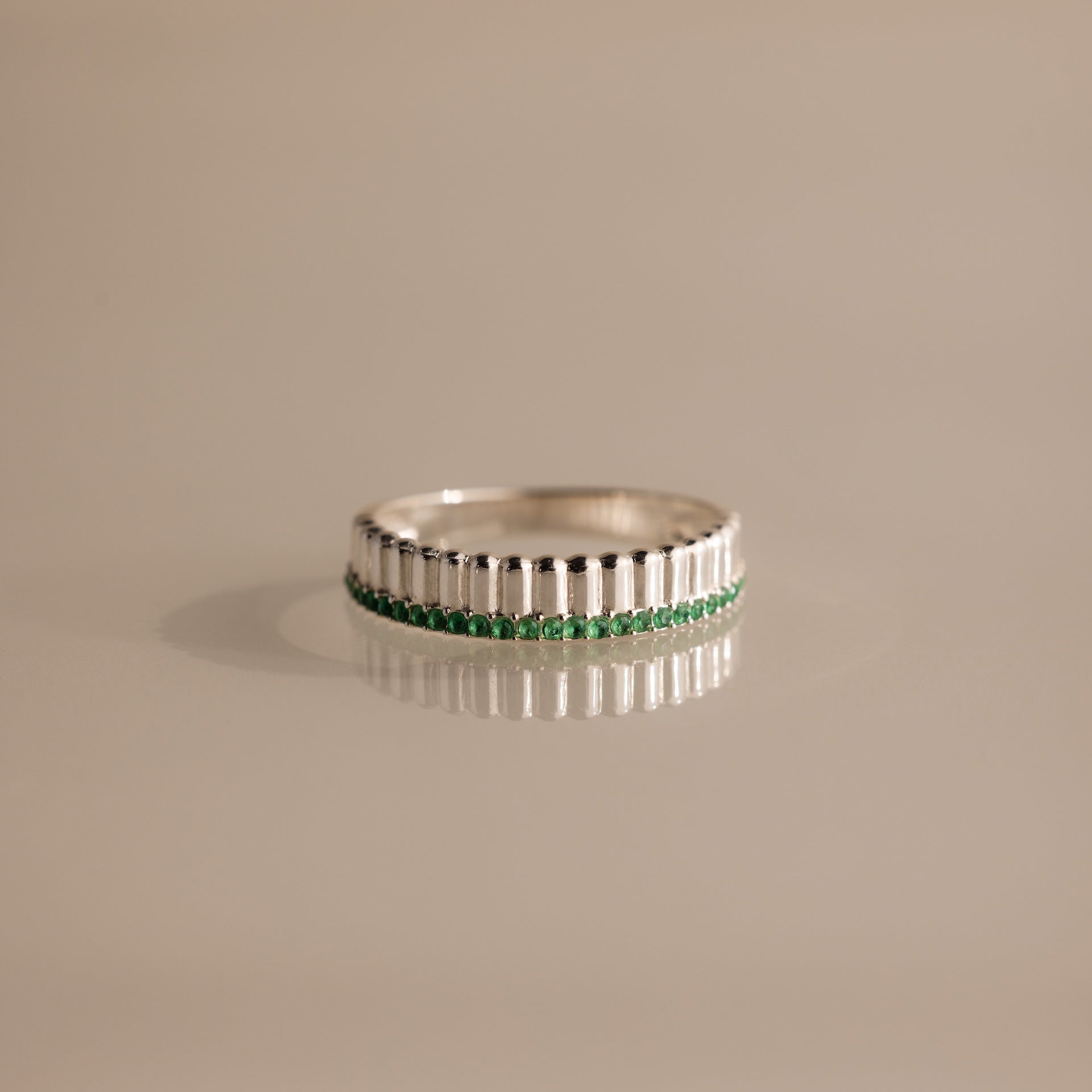 The Ribbed Emerald Stack Ring features a sculpted ridged texture and a central band of small green emeralds, ideal for everyday ring stacking. Shown on a beige surface.