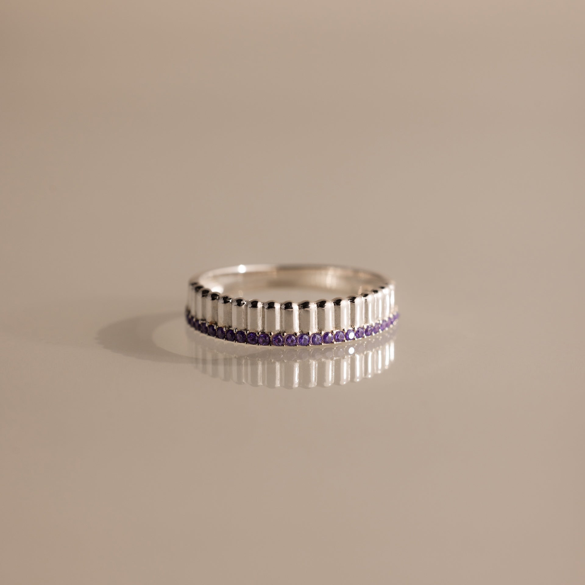 The Ribbed Amethyst Stack Ring, featuring a silver ribbed texture and a row of small purple stones, is shown on a reflective beige surface. This stylish birthstone ring is perfect for stacking and layering.