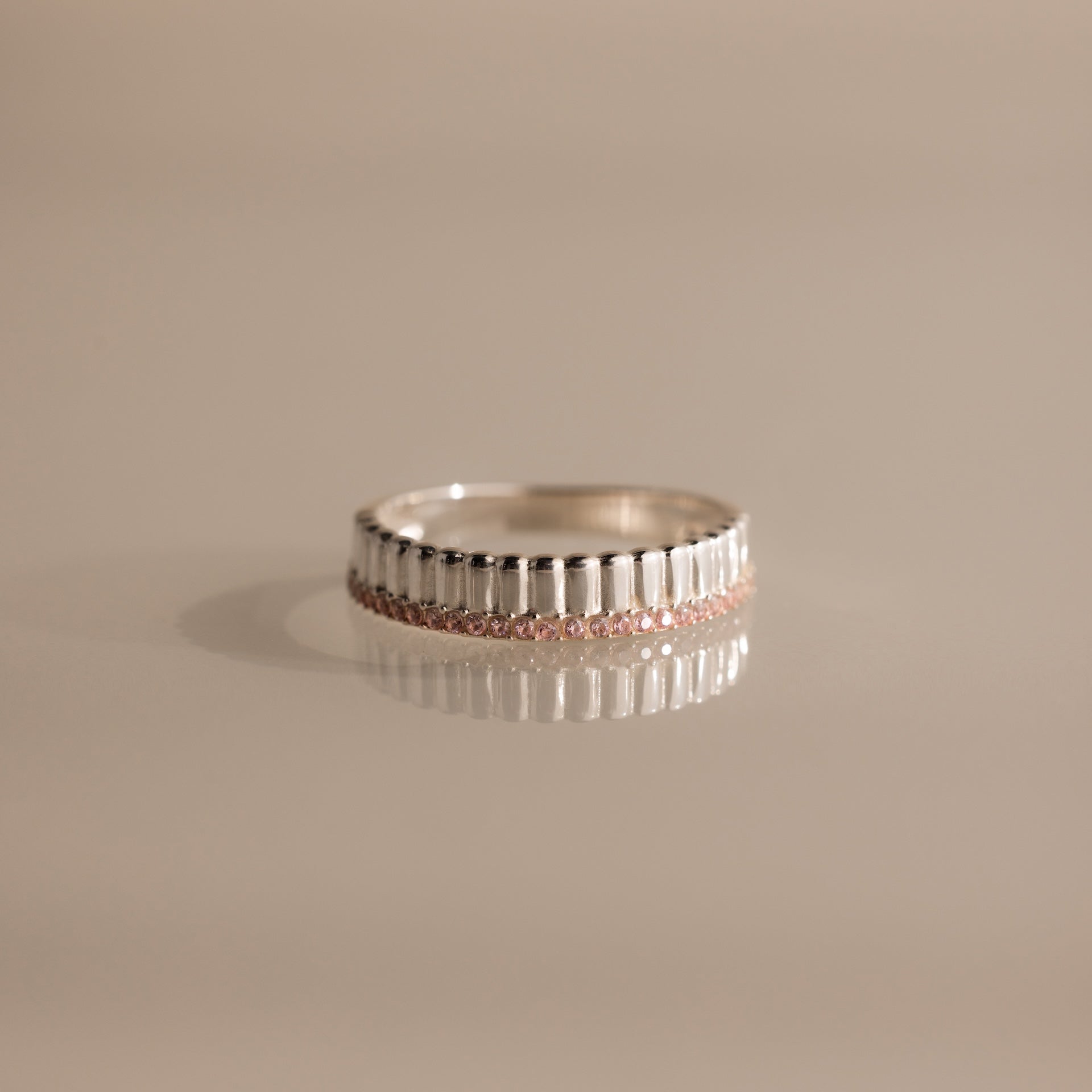 The Ribbed Pink Tourmaline Stack Ring features a sculpted ribbed texture and a row of small pink tourmaline gemstones, shown on a reflective surface. Ideal for creating your own unique ring stack style.