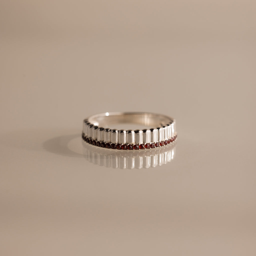 The Ribbed Garnet Stack Ring features a row of small red garnet stones set in silver, ideal for layering with other rings and shown on a beige surface.