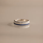 The Ribbed Sapphire Stack Ring is a modern piece with sculpted ribbed texture, vertical ridges, and a row of small blue sapphires set around the band, displayed on a beige background.