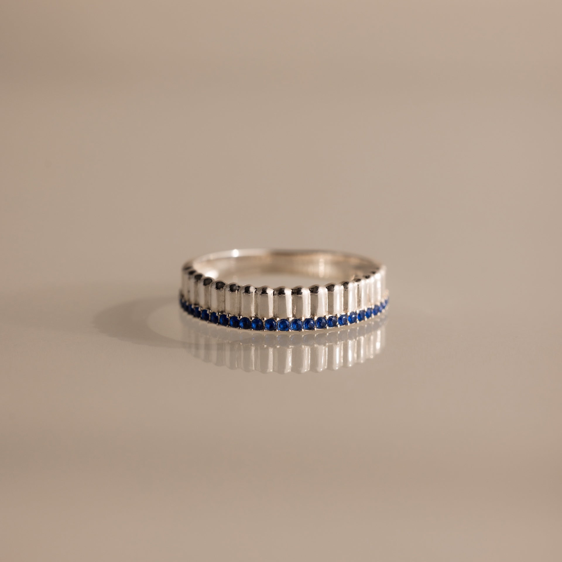 The Ribbed Sapphire Stack Ring is a modern piece with sculpted ribbed texture, vertical ridges, and a row of small blue sapphires set around the band, displayed on a beige background.