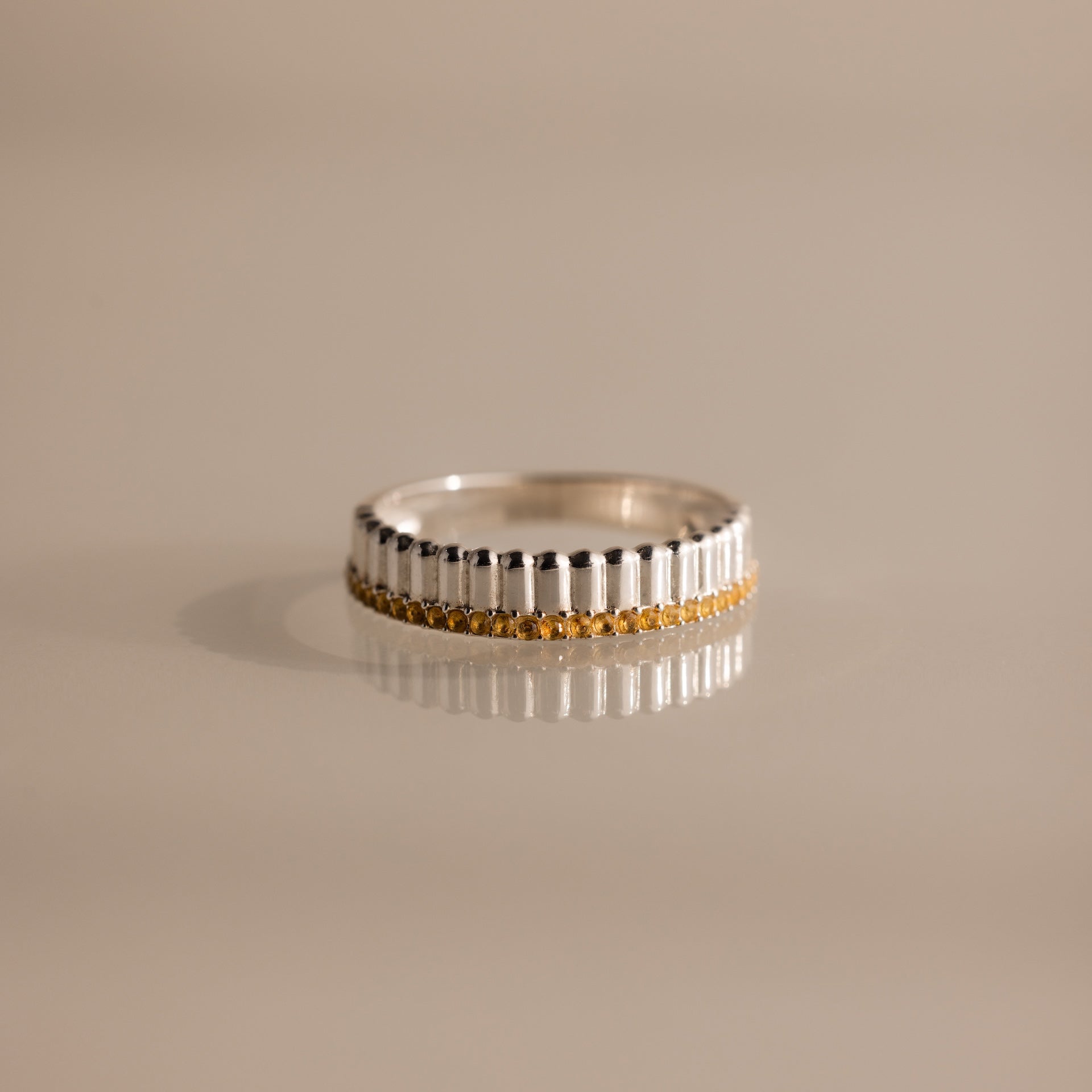 The Ribbed Topaz Stack Ring features a sculpted ribbed texture and a row of small yellow topaz stones set around the band, displayed on a shiny surface.