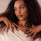 A woman with curly hair poses with her hands near her face and chest, wearing an off-shoulder top and the Ribbed Birthstone Stack Ring.