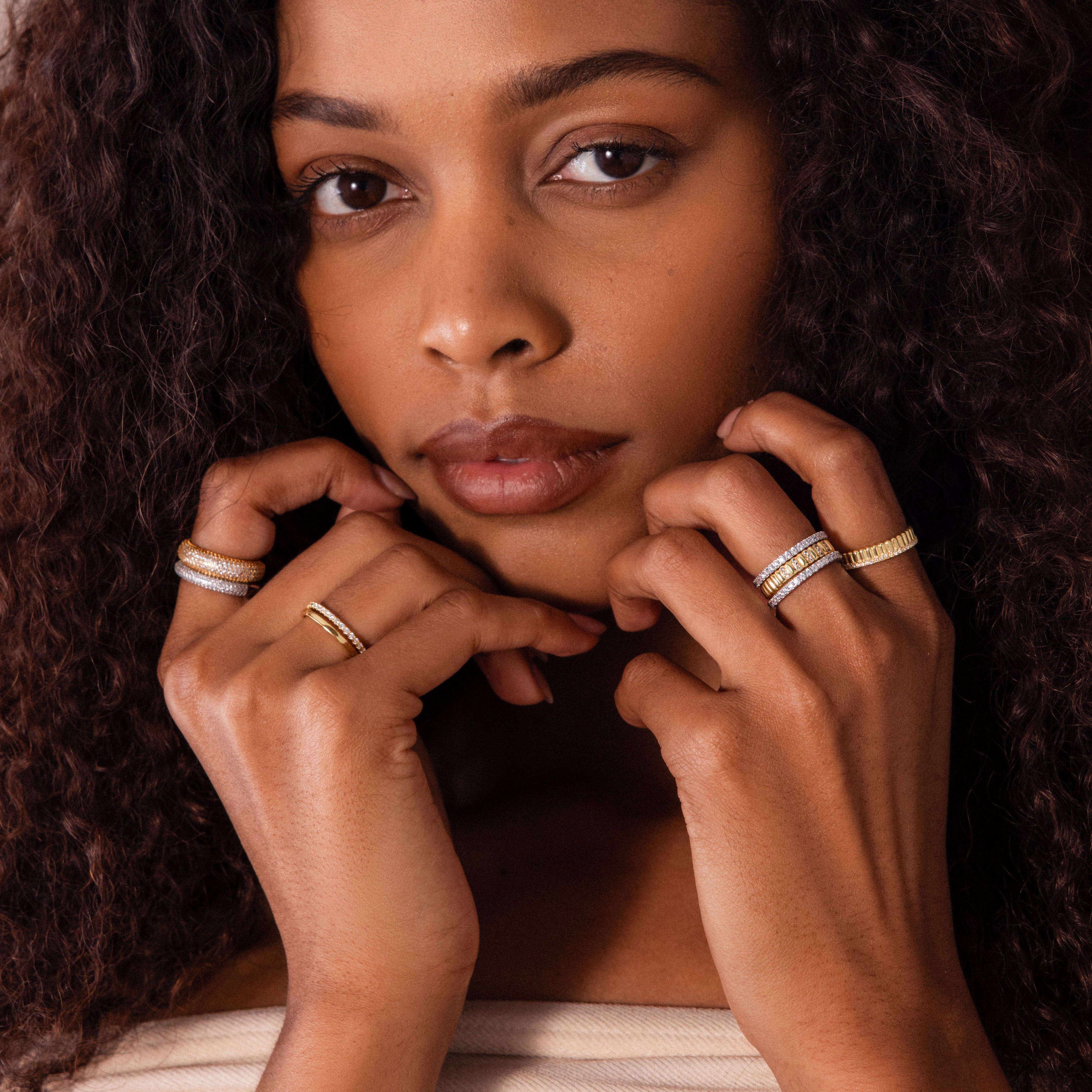 A woman with curly hair poses with her hands near her face, wearing the Ribbed Amethyst Stack Ring along with gold and silver bands to highlight the ring’s unique ribbed texture.