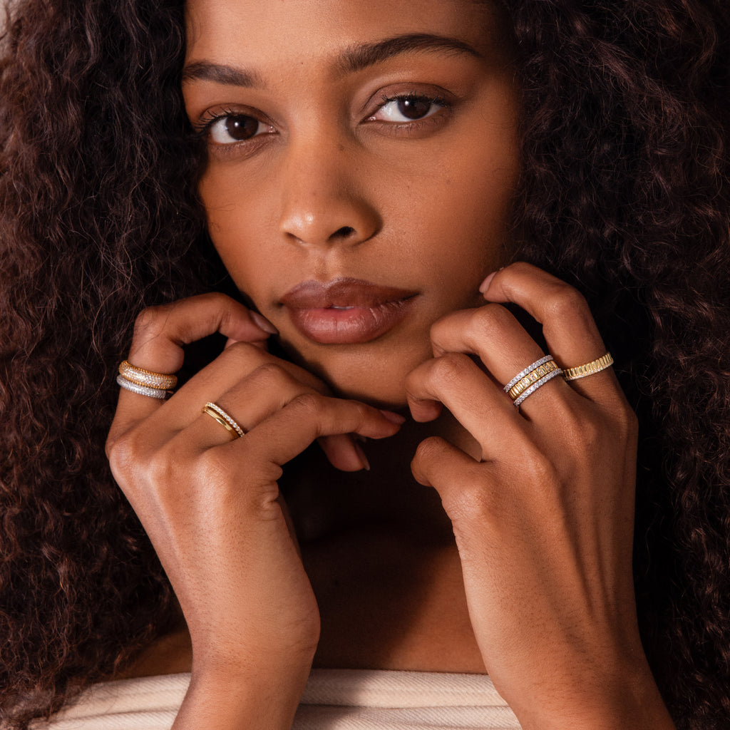 A woman with curly hair poses, wearing multiple gold and silver rings on her fingers, including a modern stackable ring and the Ribbed Aquamarine Stack Ring.