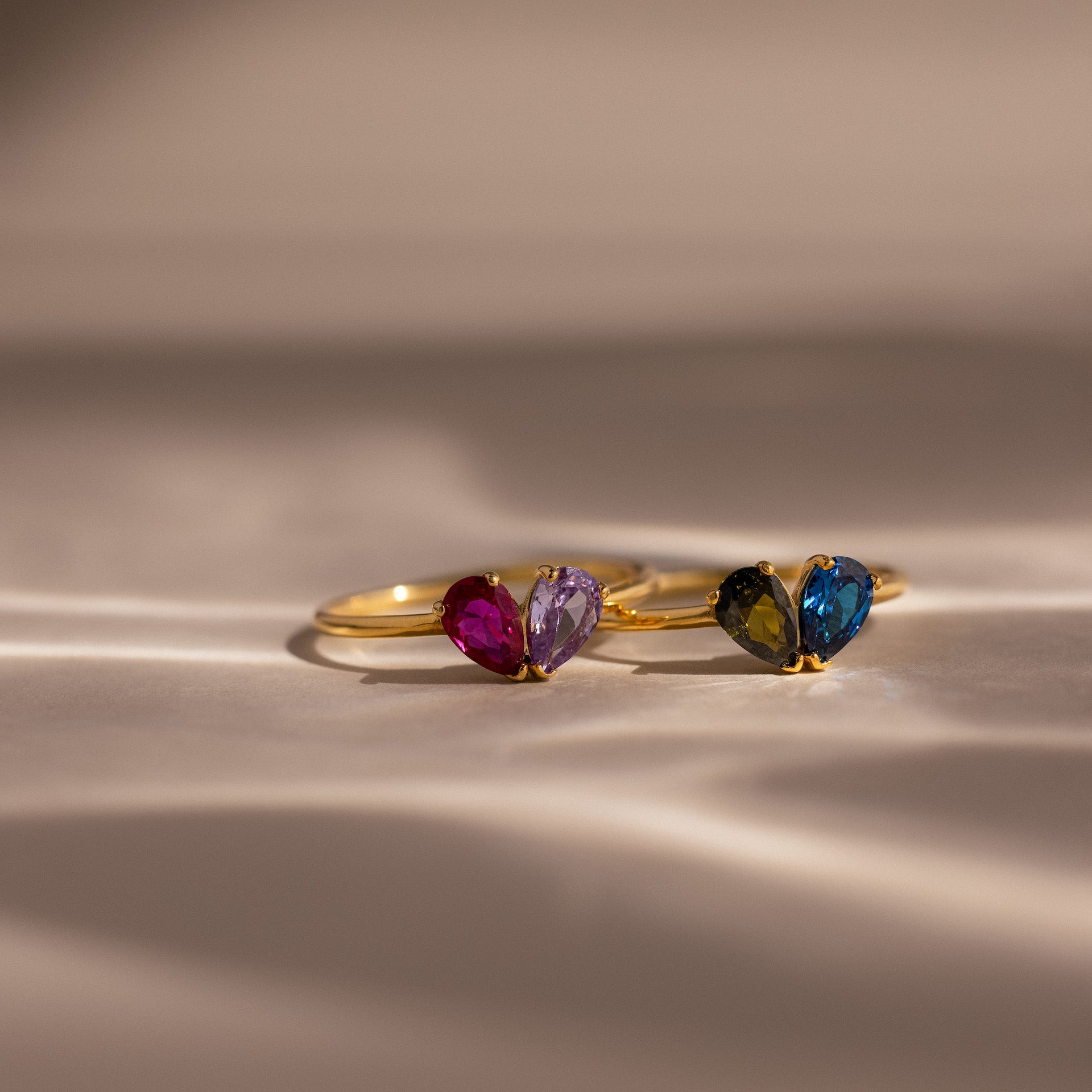Two Toi et Moi Teardrop Heart Rings, each with two heart-shaped gemstones in vivid colors, are elegantly displayed on a soft beige surface.