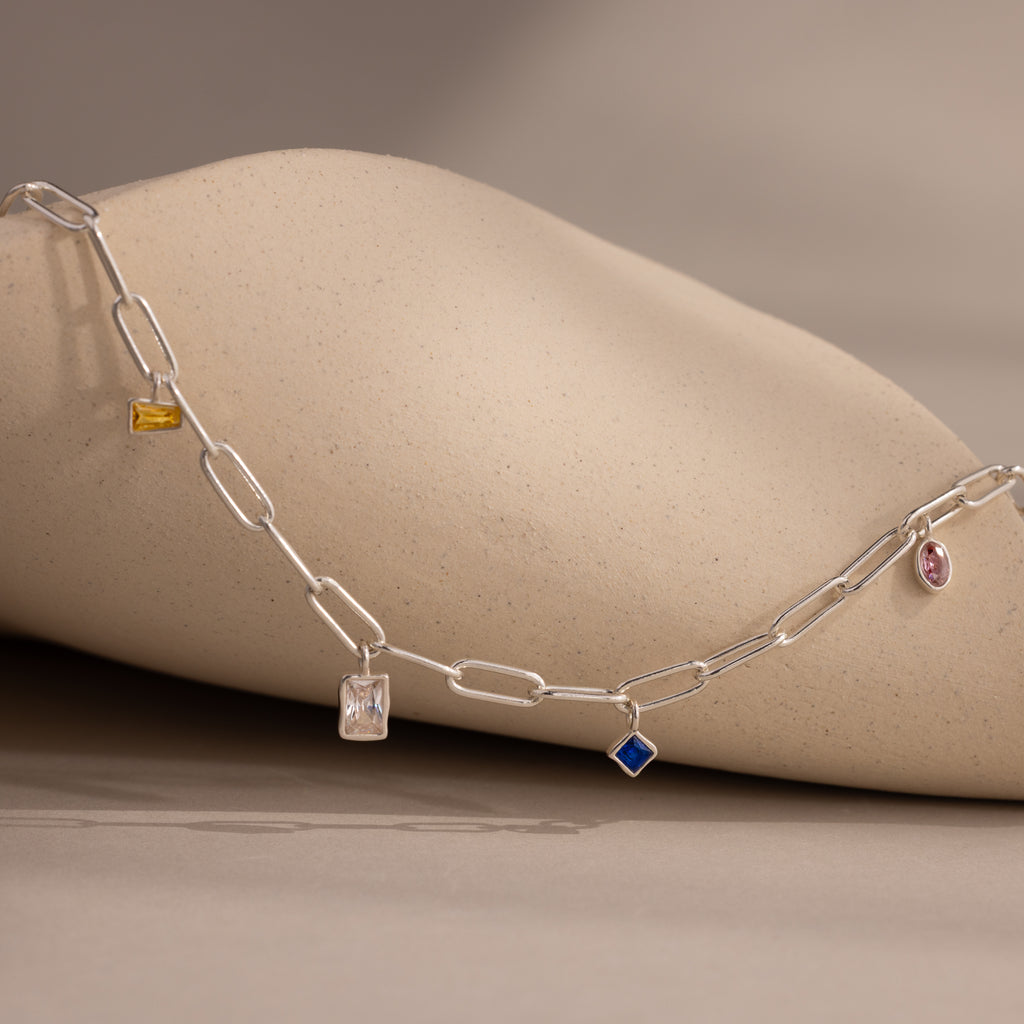 The Mixed Birthstone Charm Necklace features colorful, unique charms on a silver chain, displayed on a beige textured surface—ideal for anyone who loves personalized jewelry.