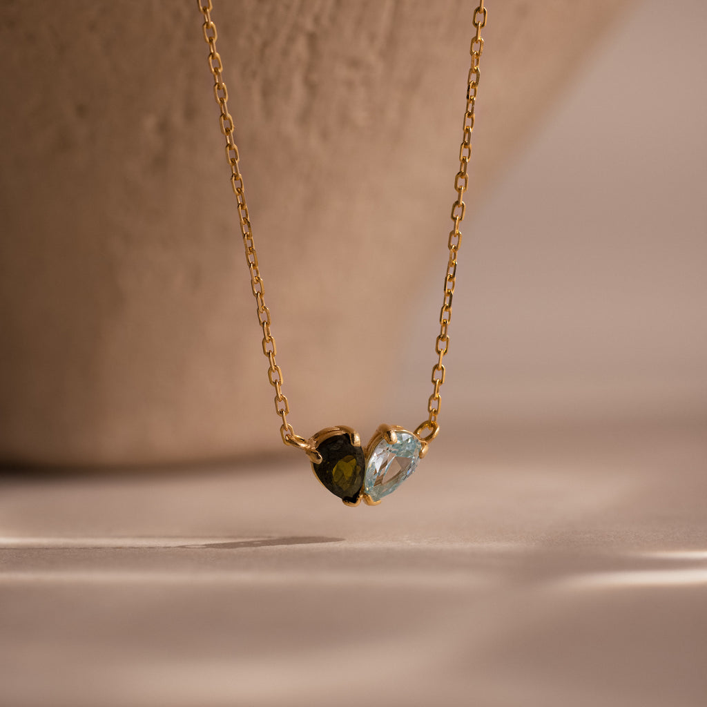The Toi et Moi Teardrop Heart Necklace in 18K Gold showcases two small heart-shaped stones—one dark, one light—gracefully set against a neutral background.