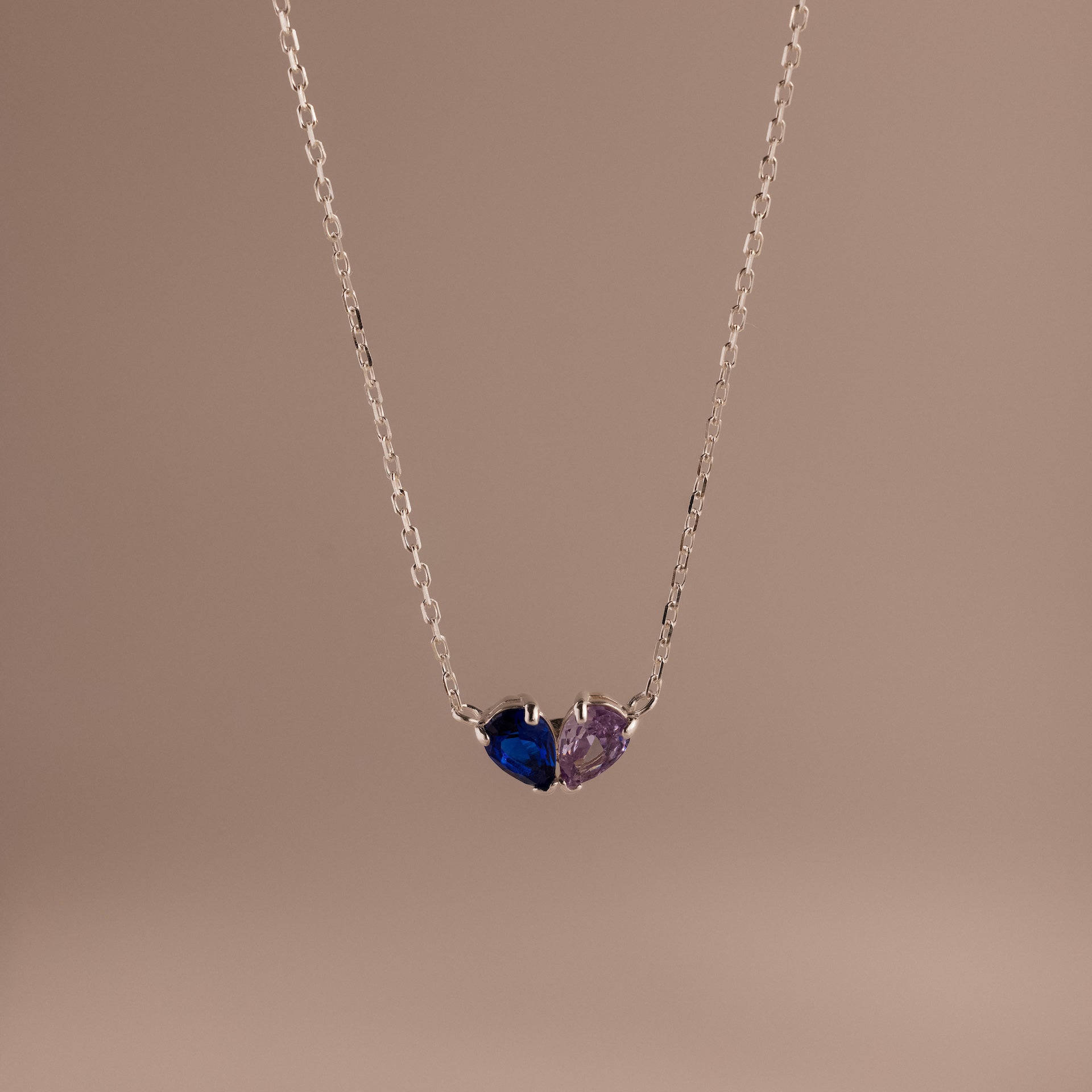 The Toi et Moi Teardrop Heart Necklace in Sterling Silver features two pear-cut stones—one blue, one purple—set on a plain background; a timeless keepsake ideal for any occasion.