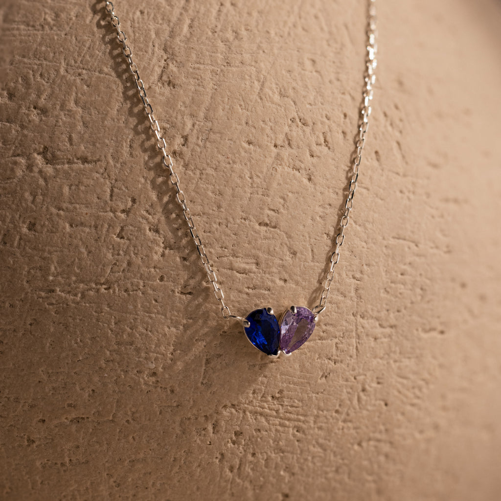 The Toi et Moi Teardrop Heart Necklace features pear-cut blue and purple stones in a silver heart design, beautifully displayed on a textured beige surface.