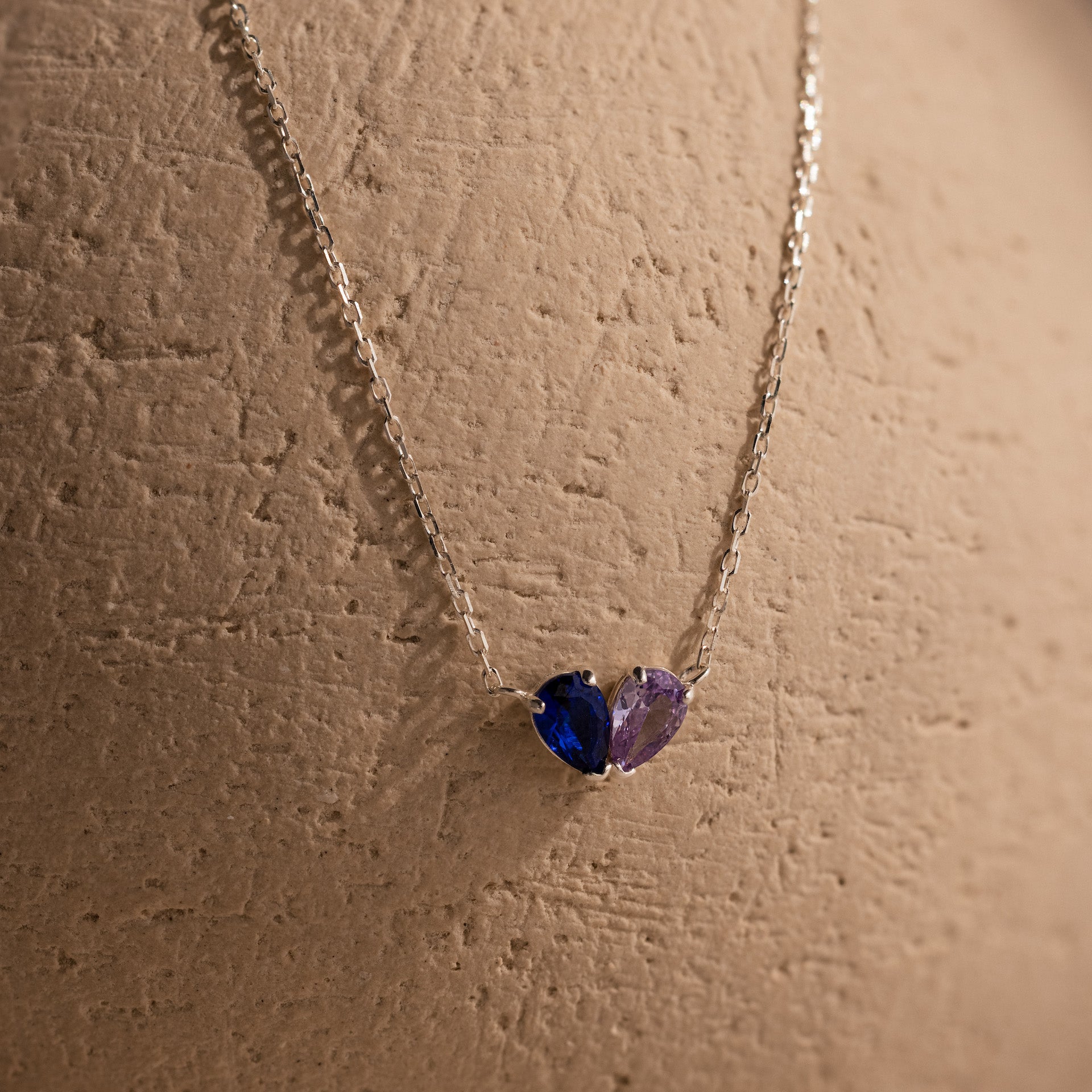 The Toi et Moi Teardrop Heart Necklace features pear-cut blue and purple stones in a silver heart design, beautifully displayed on a textured beige surface.