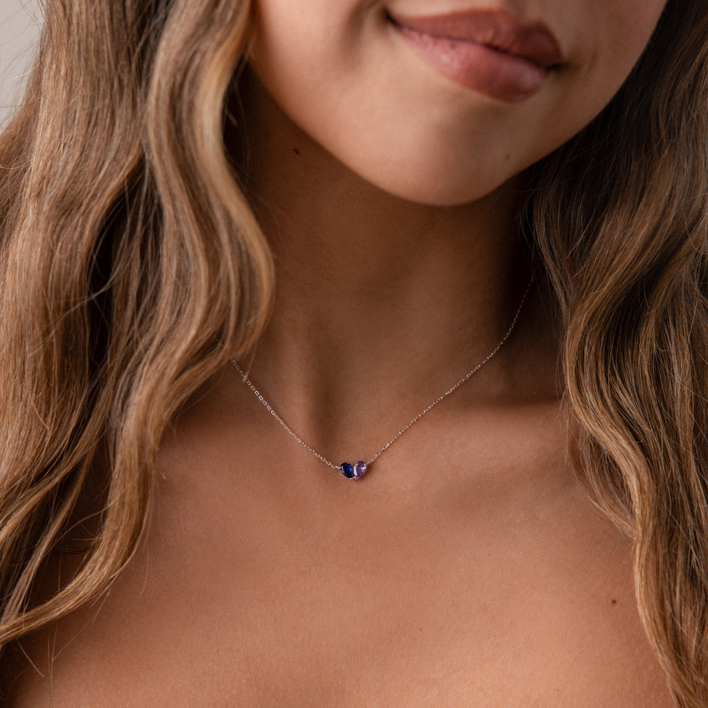 A woman with wavy hair wears the Toi et Moi Teardrop Heart Necklace in Sterling Silver, adorned with three small purple and blue pear-cut stones—a timeless keepsake.