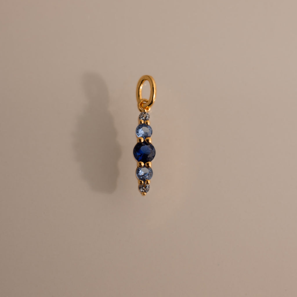 The Ombre Lace Blue Zircon Charm, a gold pendant with a vertical row of blue and silver beads, is photographed on a beige background.